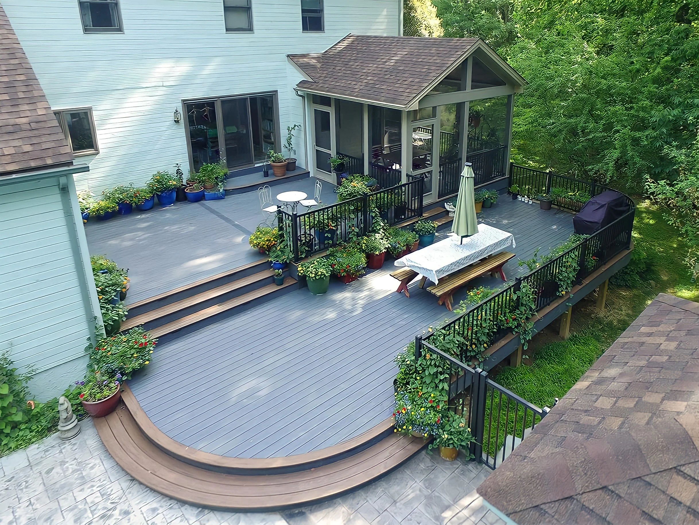 A backyard deck with potted plants, a small round table with chairs, a picnic table with an umbrella, and a grill, surrounded by trees and a house.