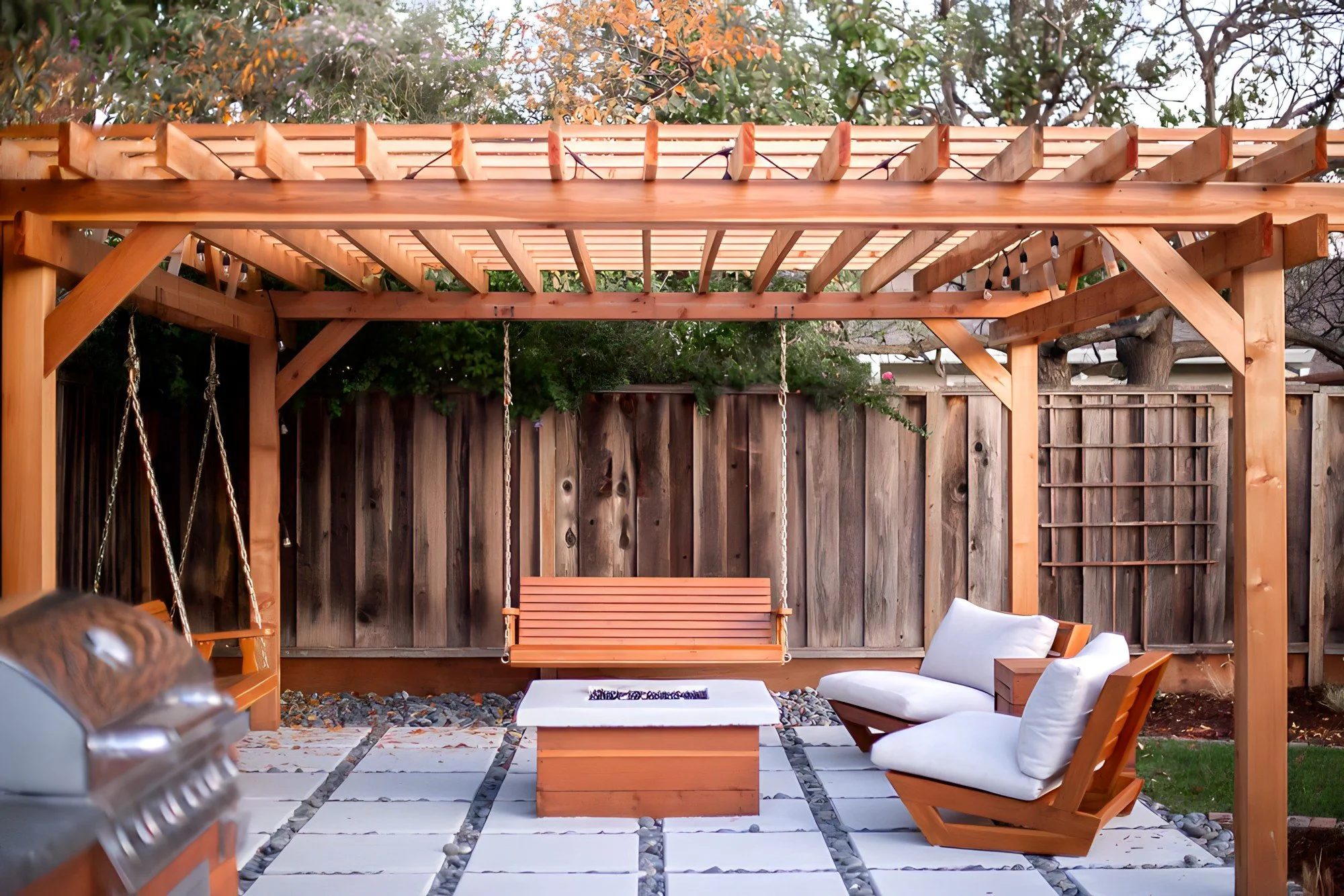 Backyard patio with wooden pergola, white cushioned chairs, a swing, a fire pit, and a barbecue grill. Wooden fence and trees in the background.