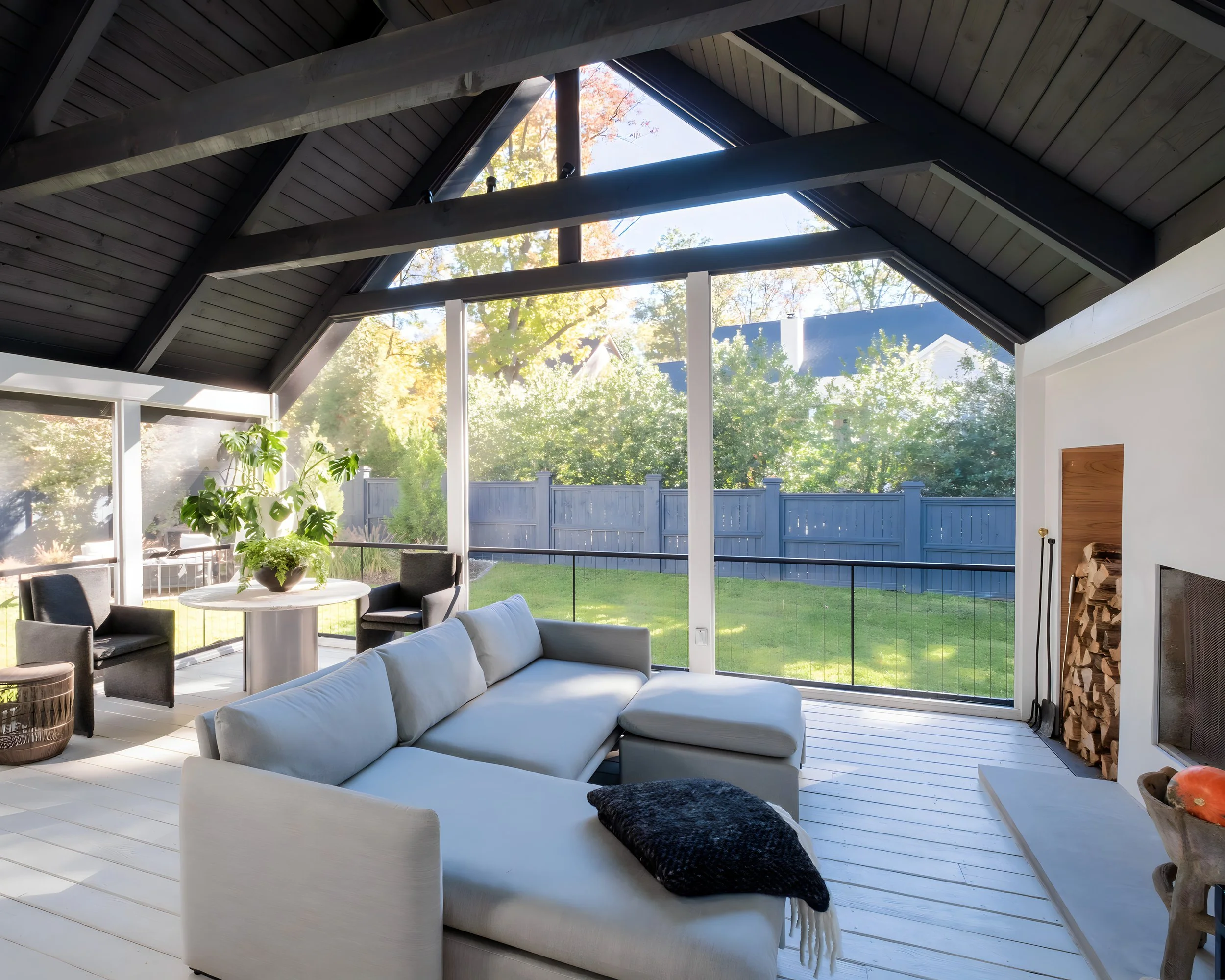 Sunlit living room with large glass windows, white sectional sofa, black armchairs, wooden ceiling with dark beams, fireplace with stacked firewood, and a view of a green backyard with trees and a blue fence.