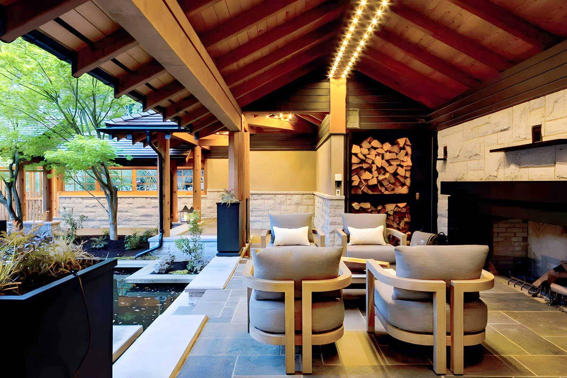  Cozy outdoor patio with seating area, stone fireplace, wood stacked for fire, and lush trees visible through open walls.