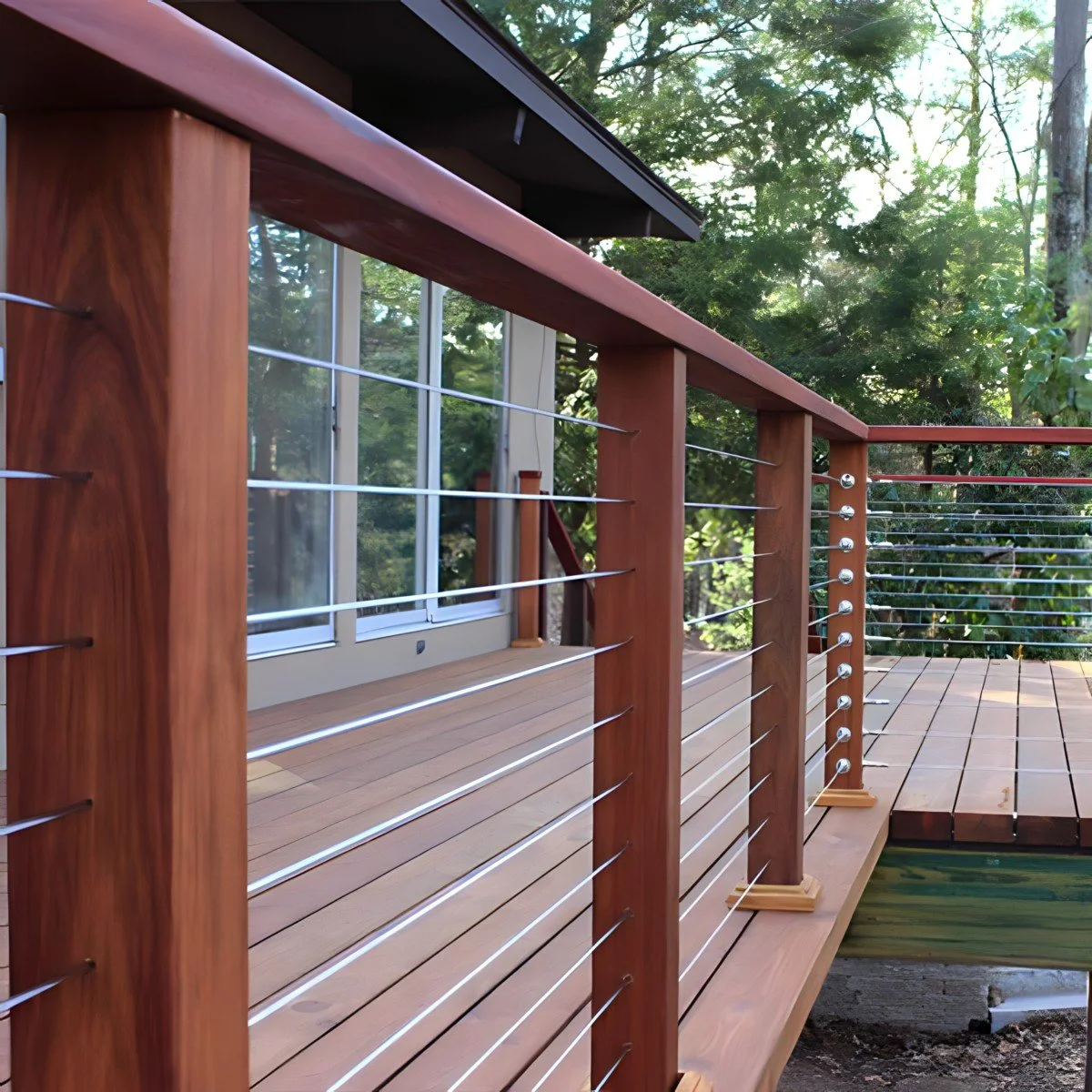 Close-up view of a wooden deck with a modern railing made of wood and metal wires, surrounded by trees and greenery.