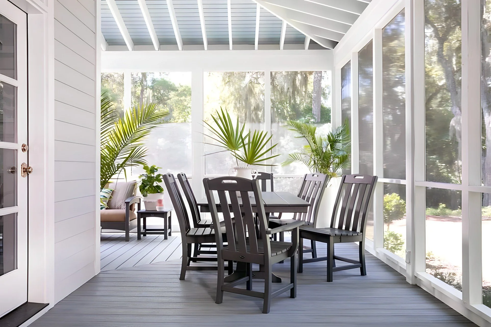 Enclosed porch with gray wooden flooring, black dining table with four matching chairs, large potted plants, and a sitting area with a bench and small table, surrounded by windows and a ceiling with exposed beams.