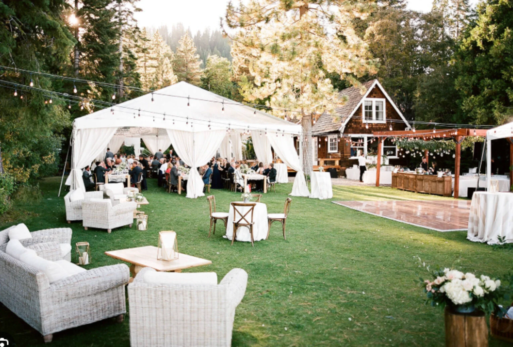 Outdoor wedding reception setup with white tent, string lights, tables, and chairs on a grassy lawn surrounded by trees and a wooden house in the background.