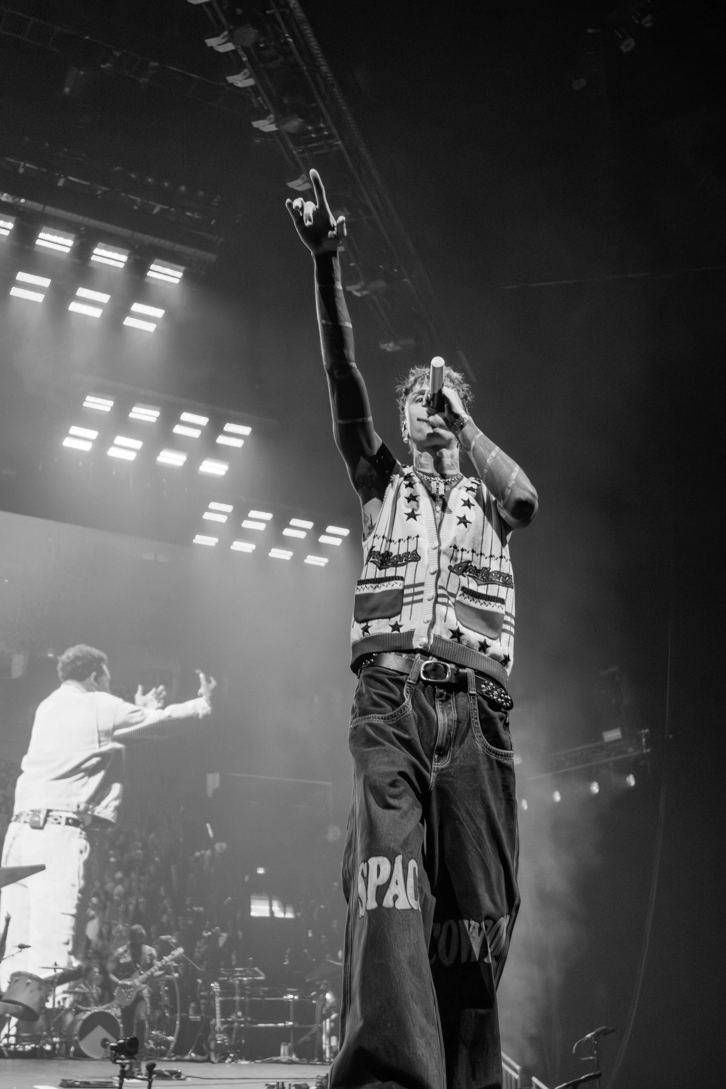 A Black and white photo of a male performer on stage holding a microphone and pointing upwards. There is a keyboard player and a drummer in the background with stage lights overhead.