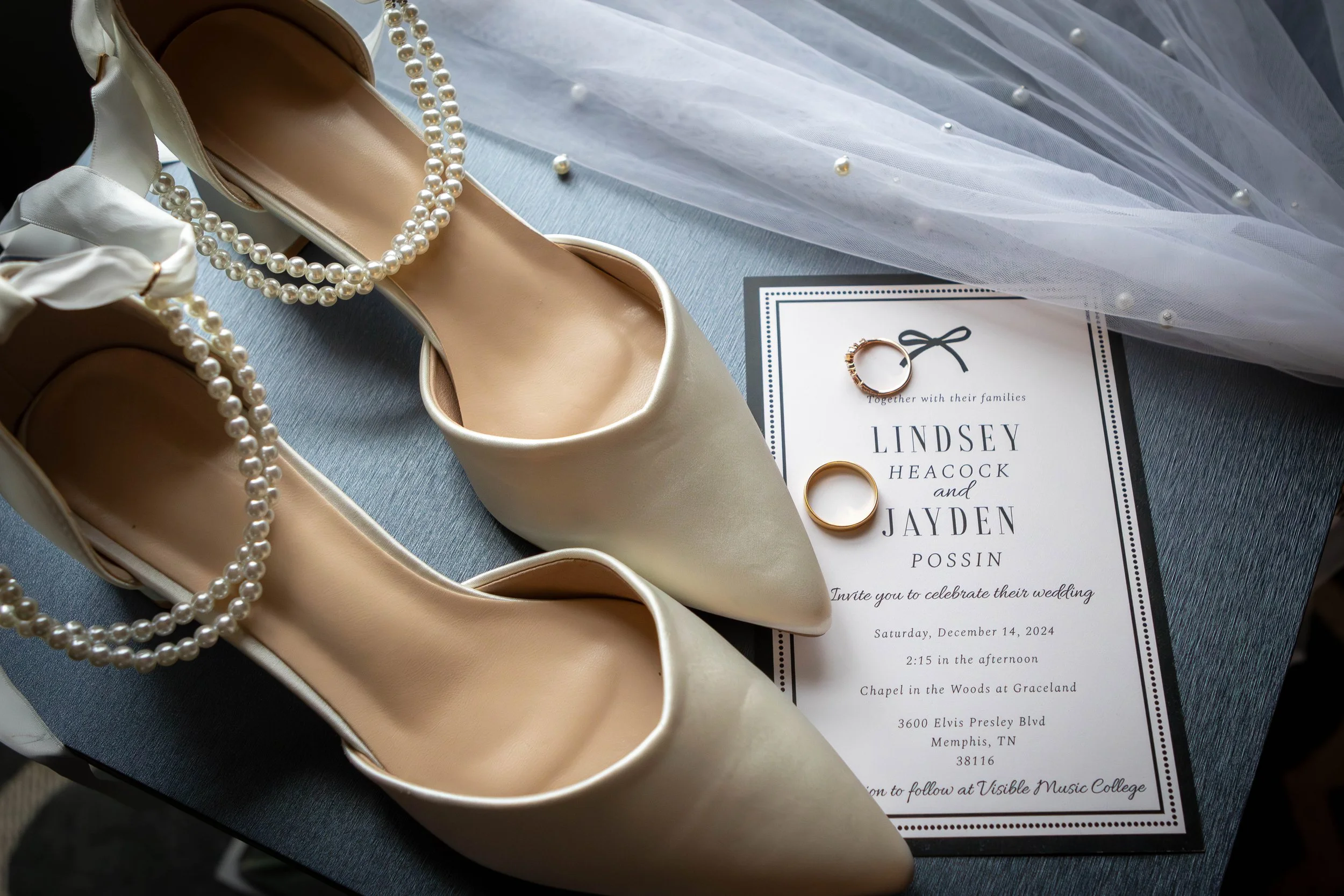 Cream-colored high heel shoes with pearl necklaces, a wedding ring, and an invitation card for a wedding celebration.