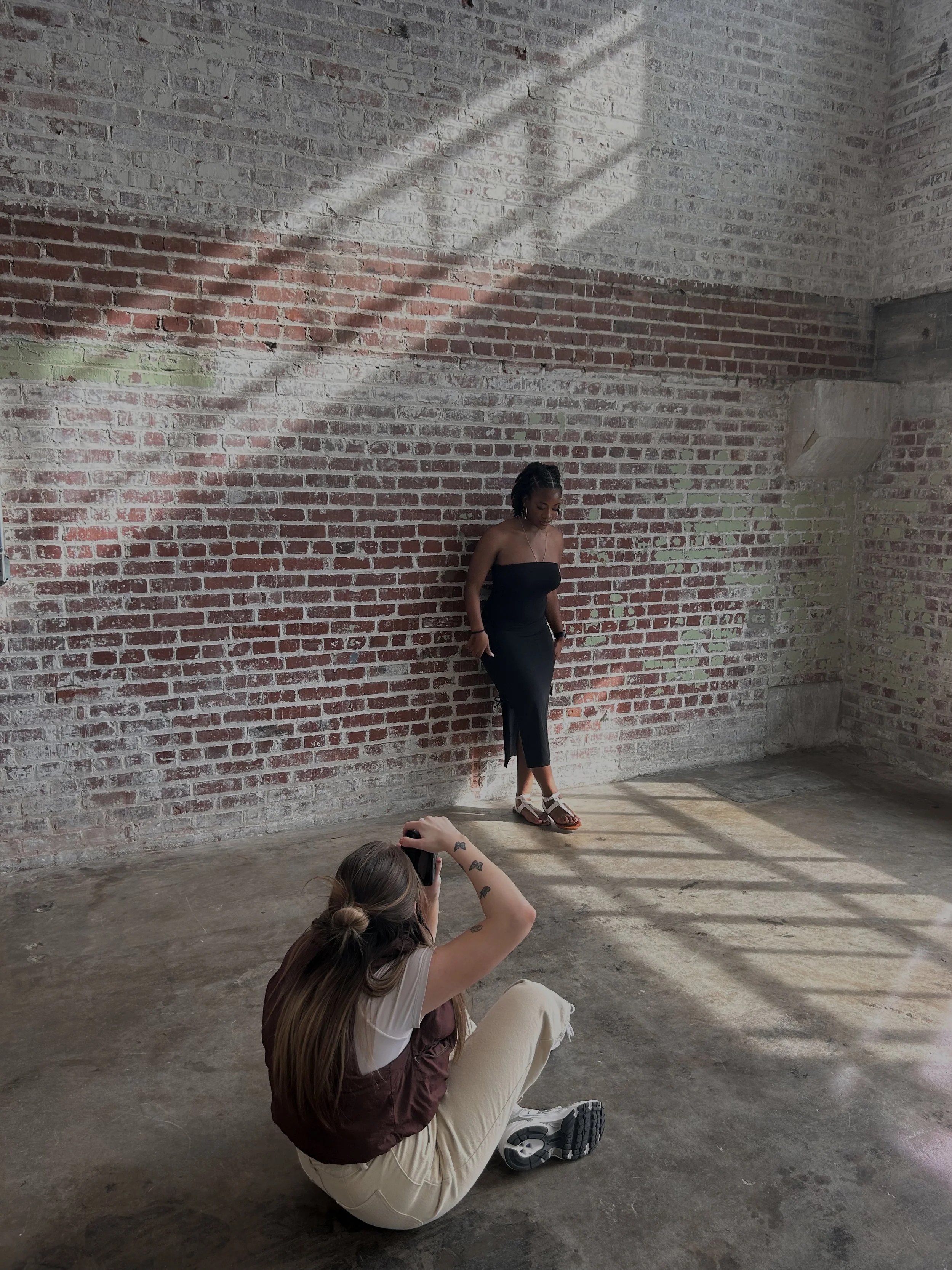 Two women in a photography shoot against an exposed brick wall, one standing and the other sitting on the floor taking her picture.