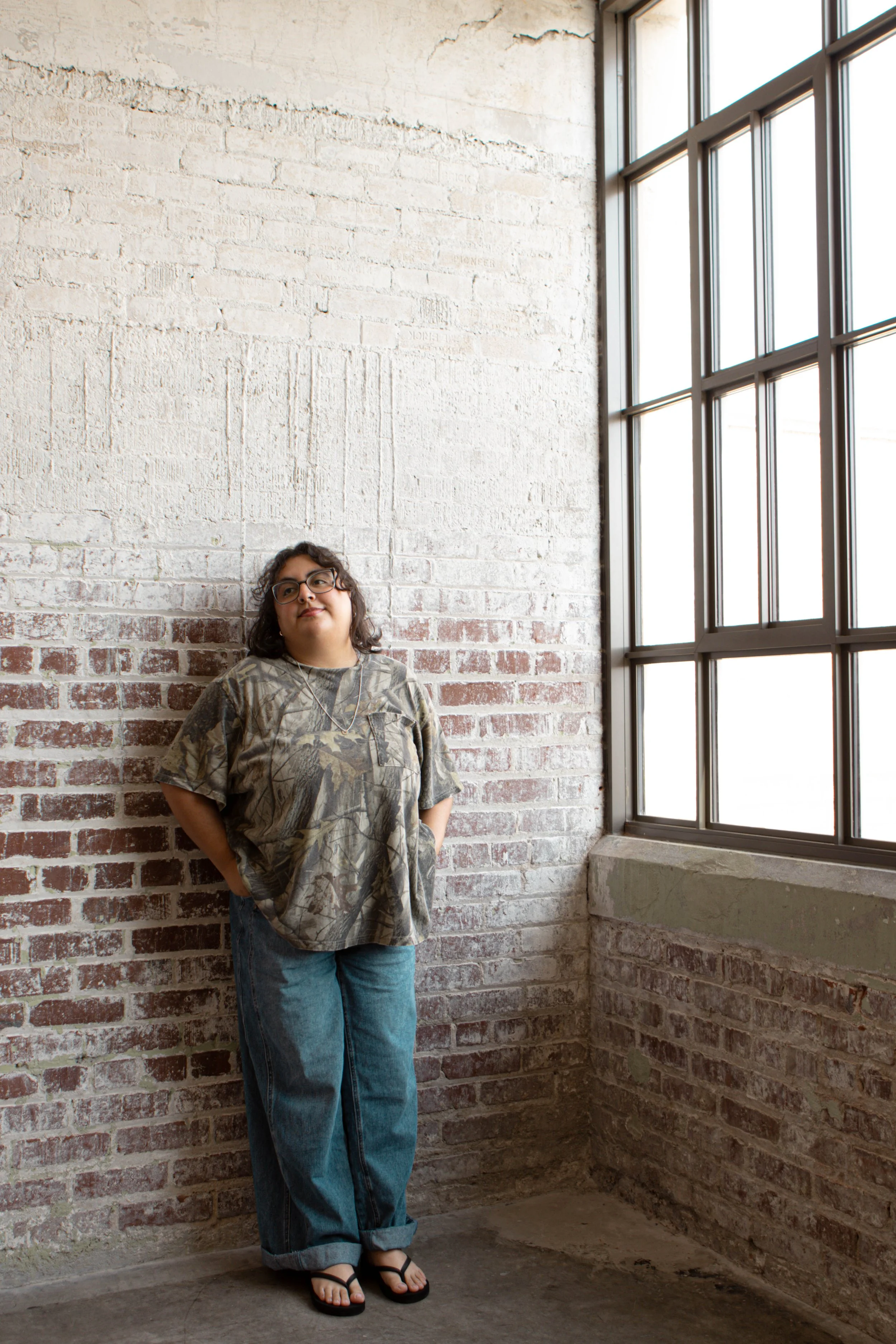 A young woman with glasses and dark curly hair stands in a corner of a room with exposed brick walls and large windows. She is wearing a camo-patterned oversized T-shirt, loose blue jeans with the cuffs rolled up, and black flip-flops, looking slight