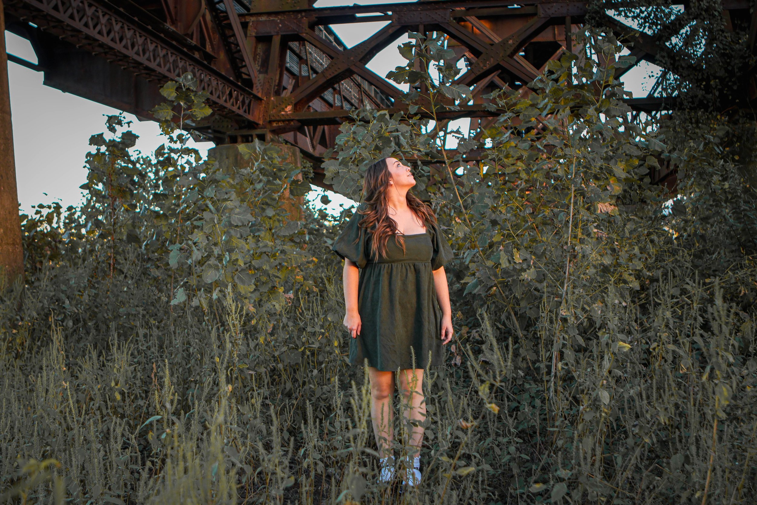 A woman standing amidst tall grass and bushes, looking up at an elevated rusted metal train bridge with industrial structure and steel beams.