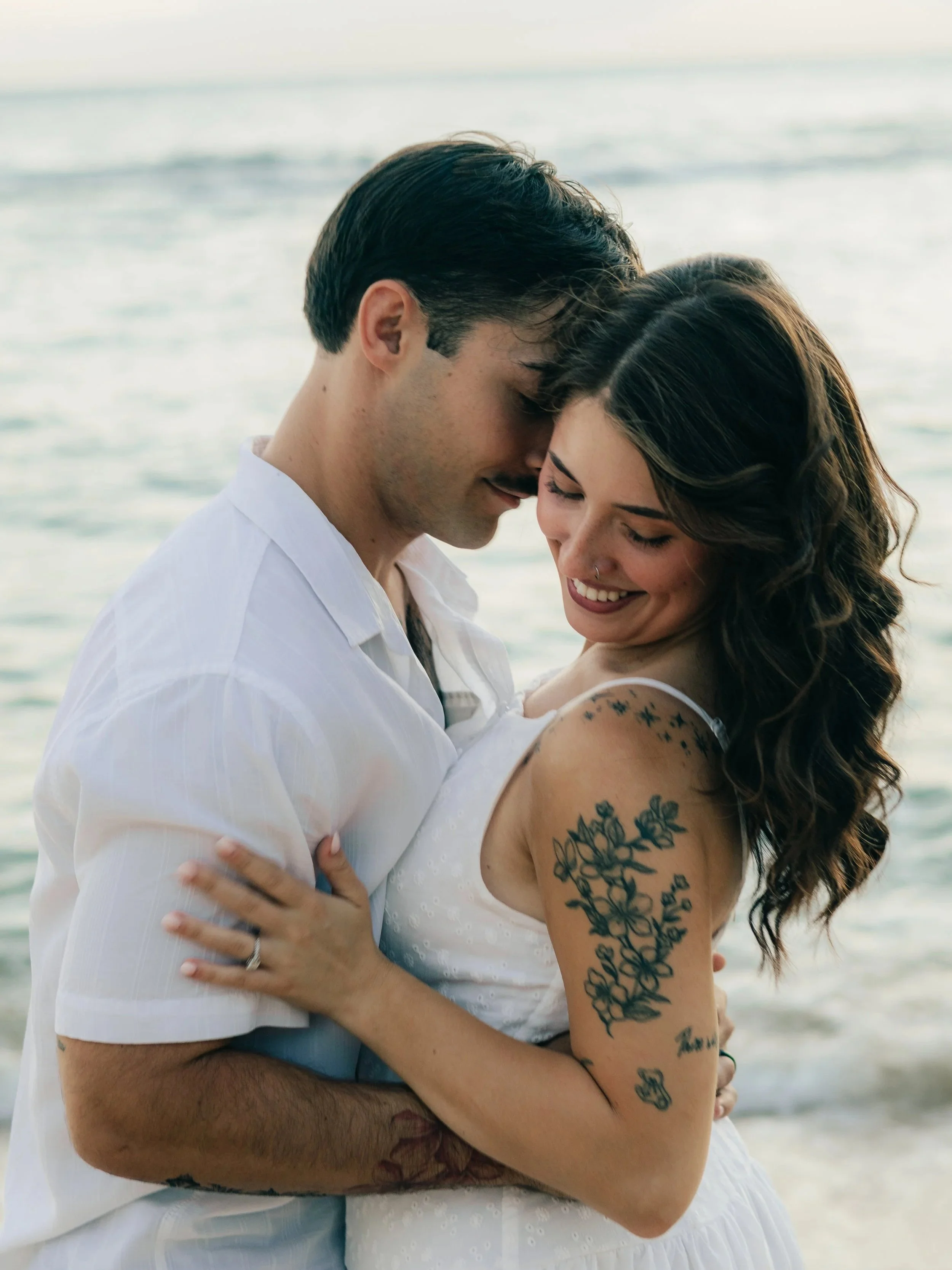 A couple embracing and smiling at each other on the beach, with the ocean in the background.