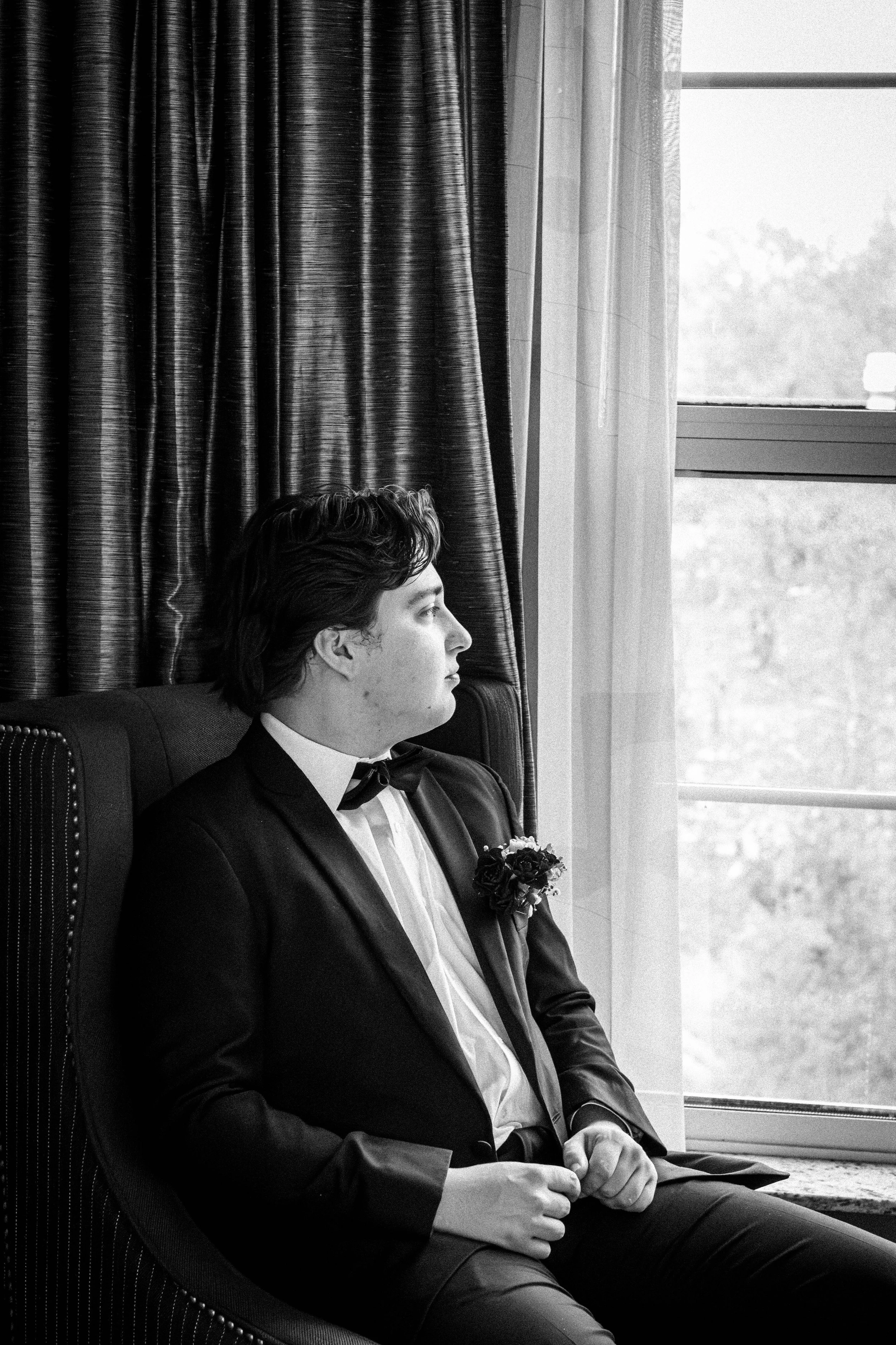 A young man with dark, wavy hair sits in a chair by a window, dressed in a formal tuxedo with a bow tie and a boutonnière. He is looking out the window with a serious expression. The room has curtains and a view of trees outside.