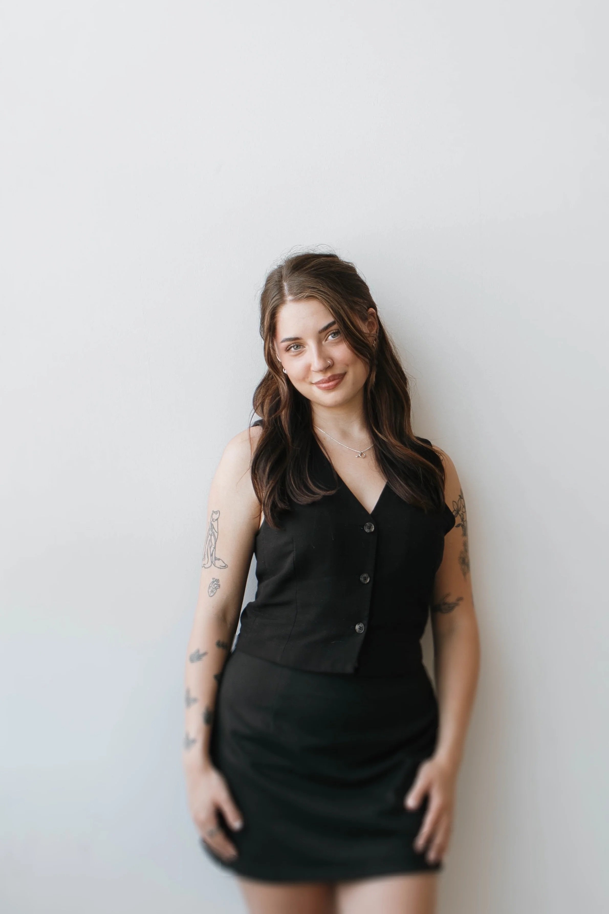 Young woman with long wavy brown hair, tattoos on arms, wearing a sleeveless black button-up dress, standing against a plain white wall.