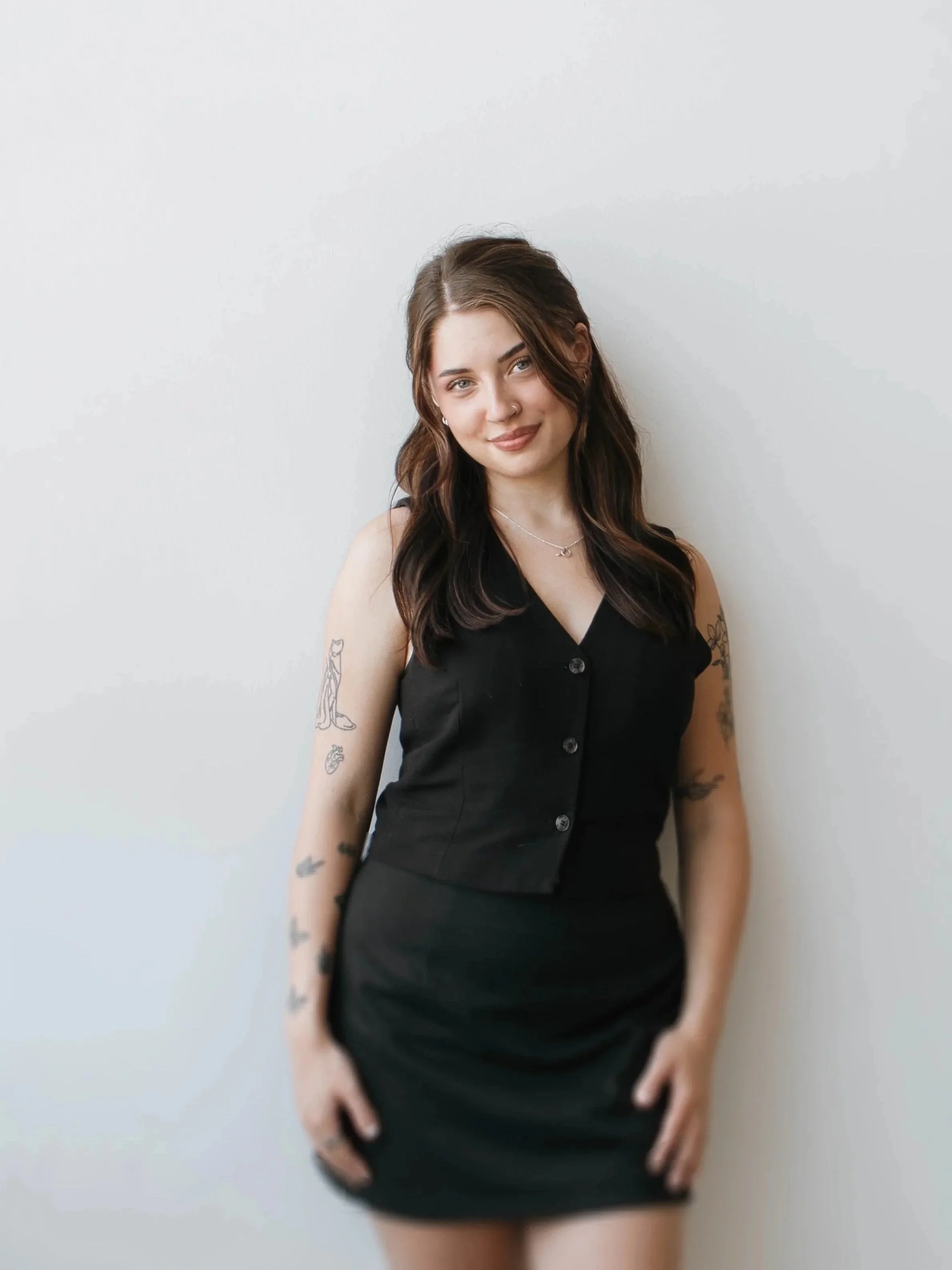 A young woman with long brown hair, wearing a black sleeveless dress with button details, standing against a plain white wall, smiling with a nose piercing and tattoos on her arms.