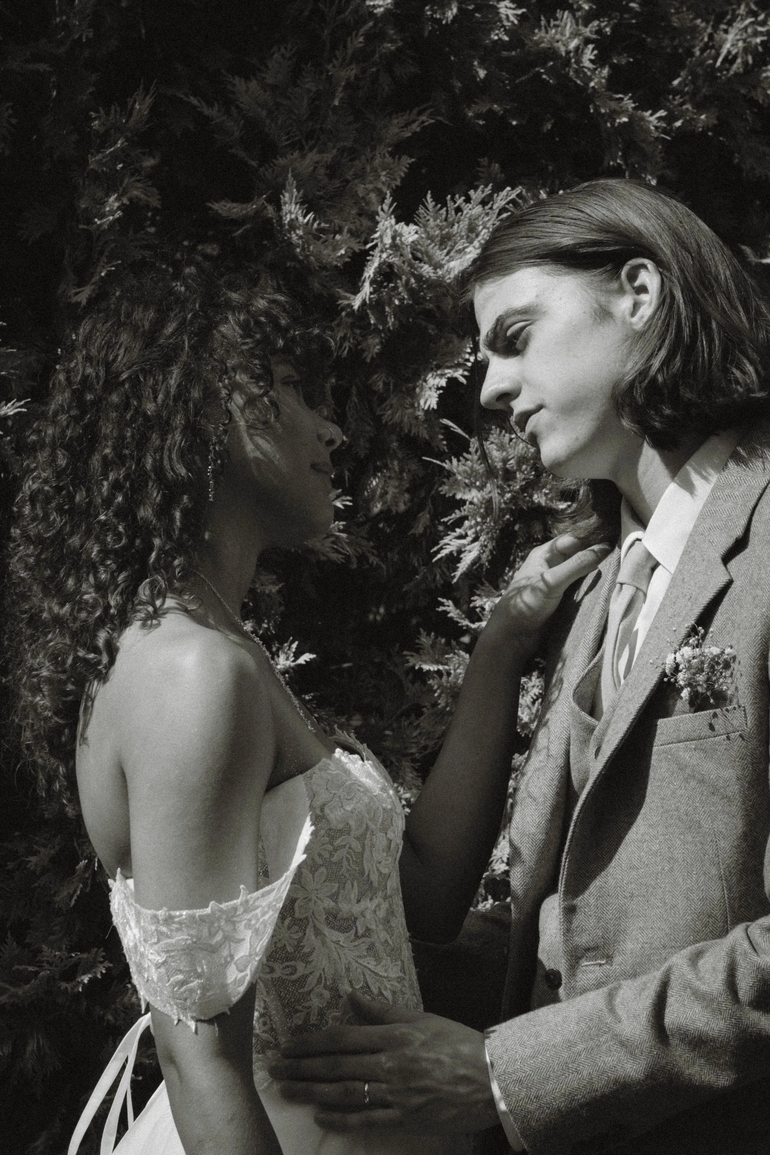 Black and white photo of a couple standing close to each other, foreheads nearly touching, surrounded by pine branches, dressed in wedding attire, in a romantic pose.