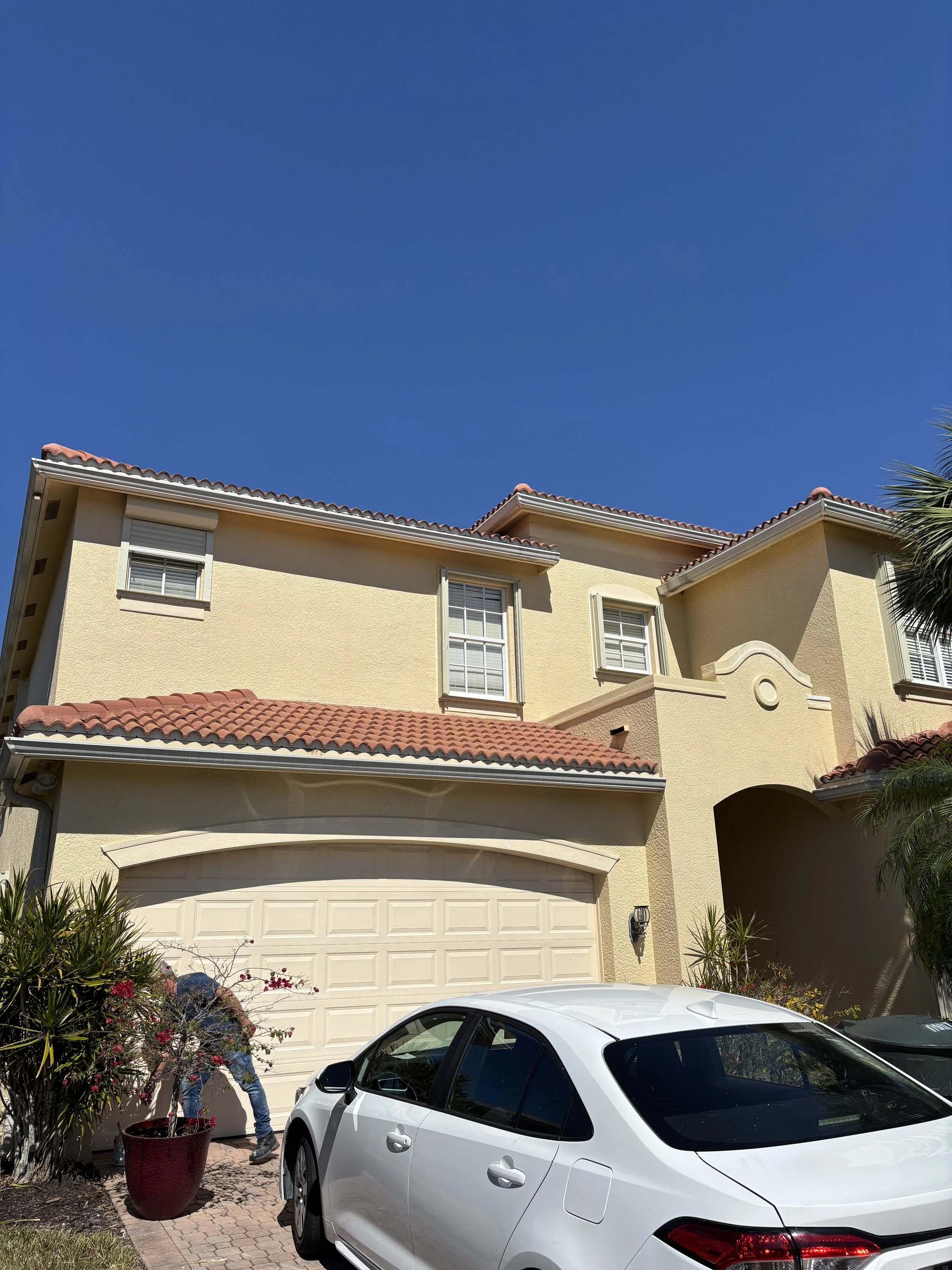A two-story yellow house with a red tiled roof, white garage door, and white-framed windows. A white car is parked in the driveway, and a person is tending to a potted plant near the garage. Clear blue sky above.