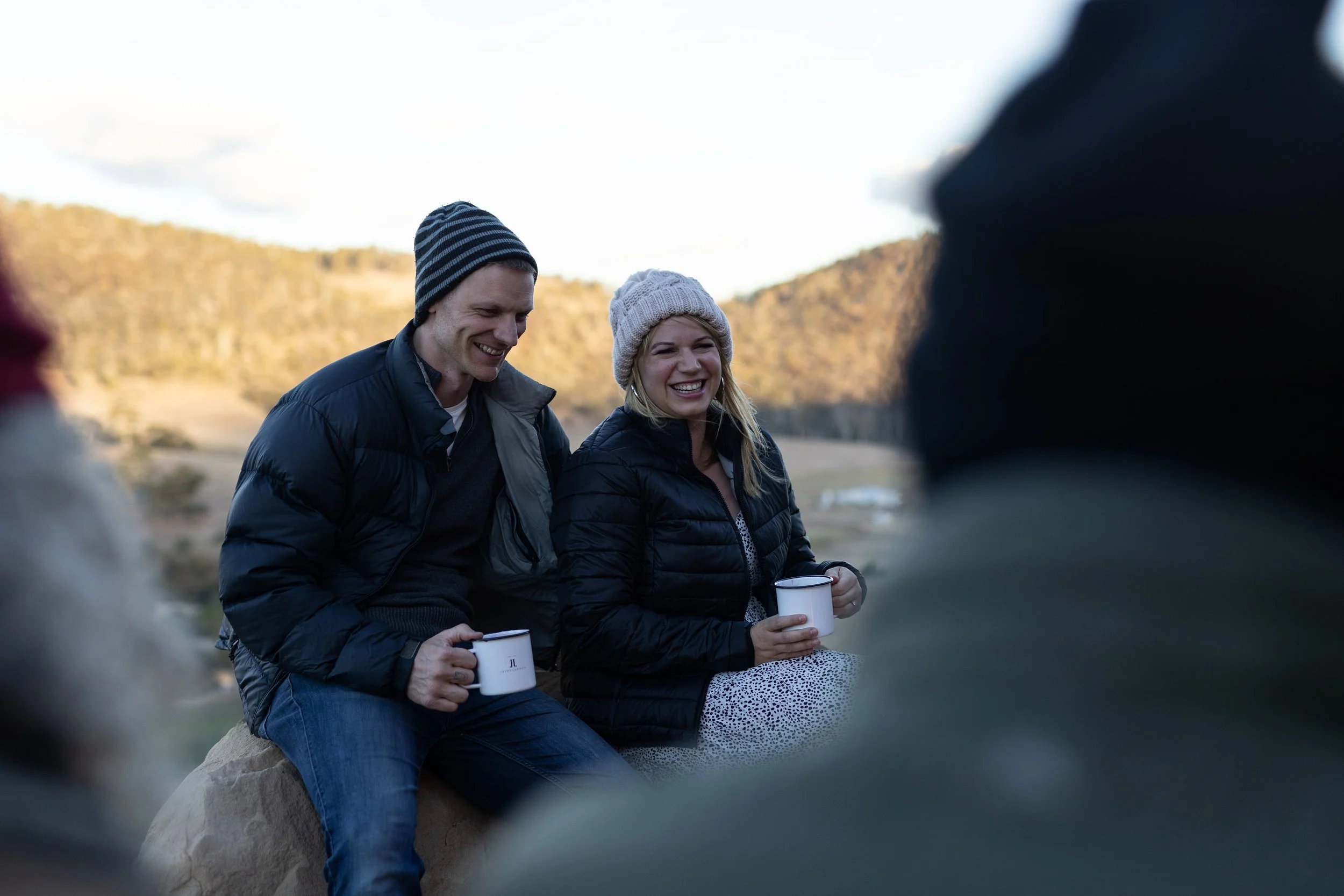 A man and woman sitting on a bench near a fire pit, enjoying hot drinks together outside on a deck with a mountain view at dusk.