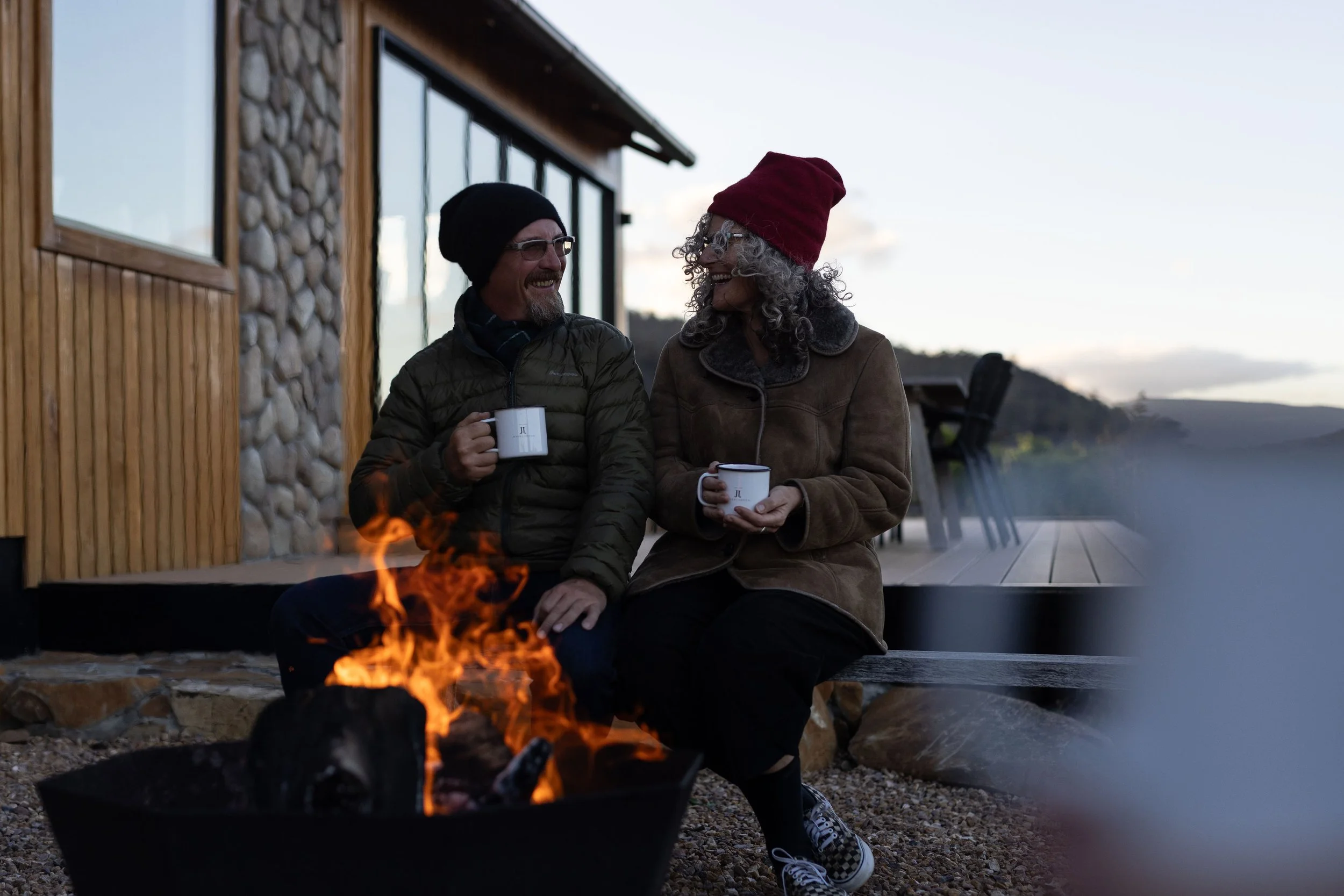 Four people sitting around a campfire and holding mugs outdoors in a hilly landscape.