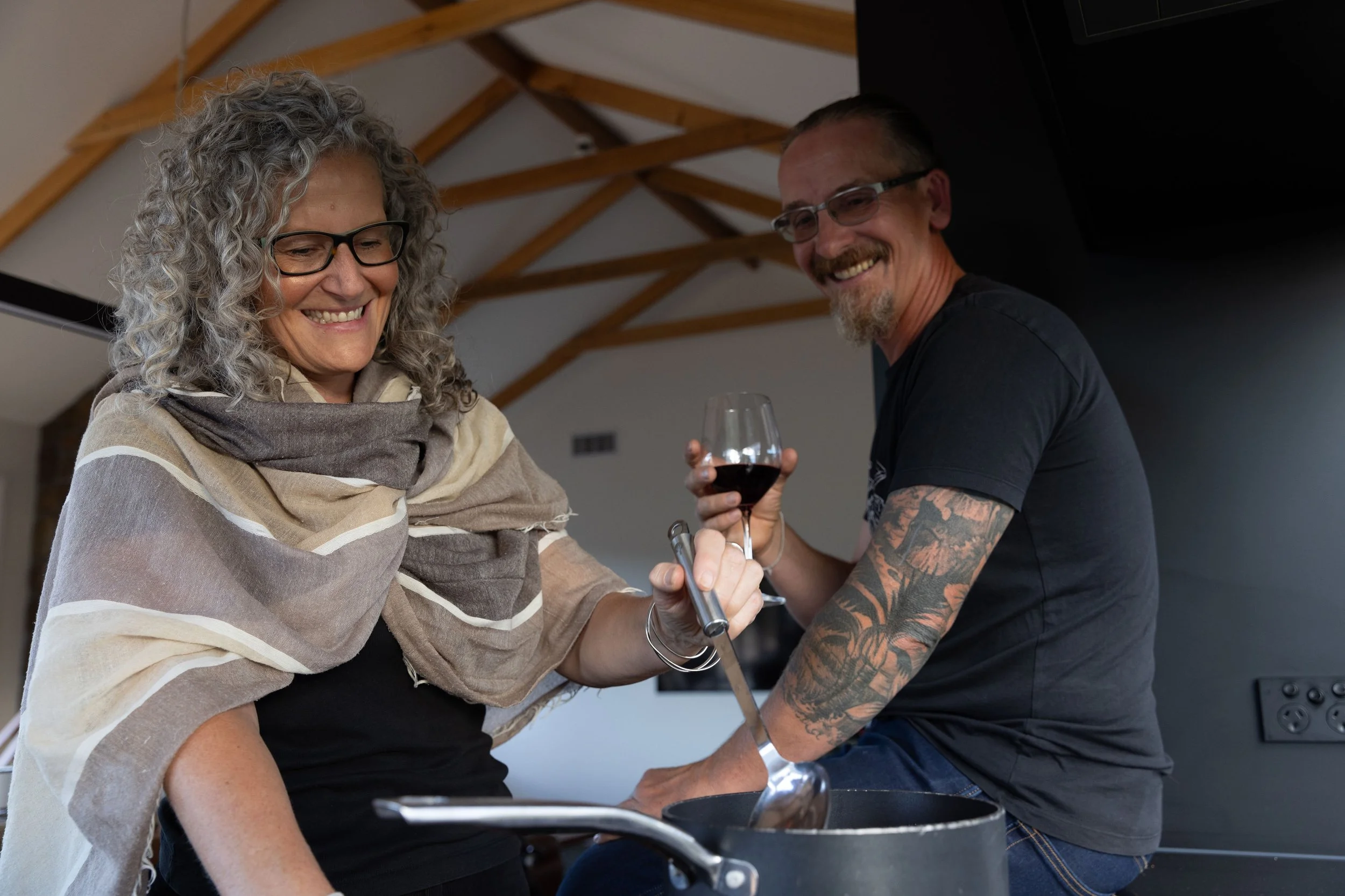 A smiling woman with curly gray hair and glasses holding a metal spatula, standing next to a smiling man with tattoos, glasses, and a beard, holding a glass of red wine in a kitchen with wooden ceiling beams.