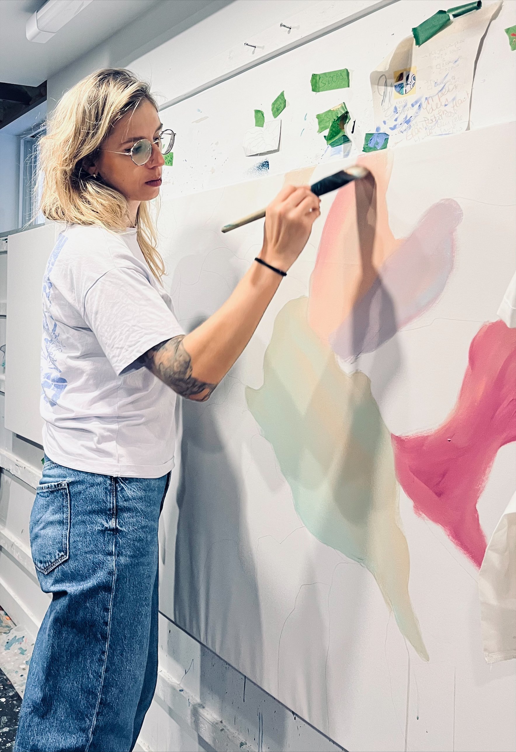 Une femme avec des lunettes et des cheveux blonds, portant un t-shirt blanc et un jean, peint sur une grande toile abstraite aux couleurs pastel, dans un atelier.