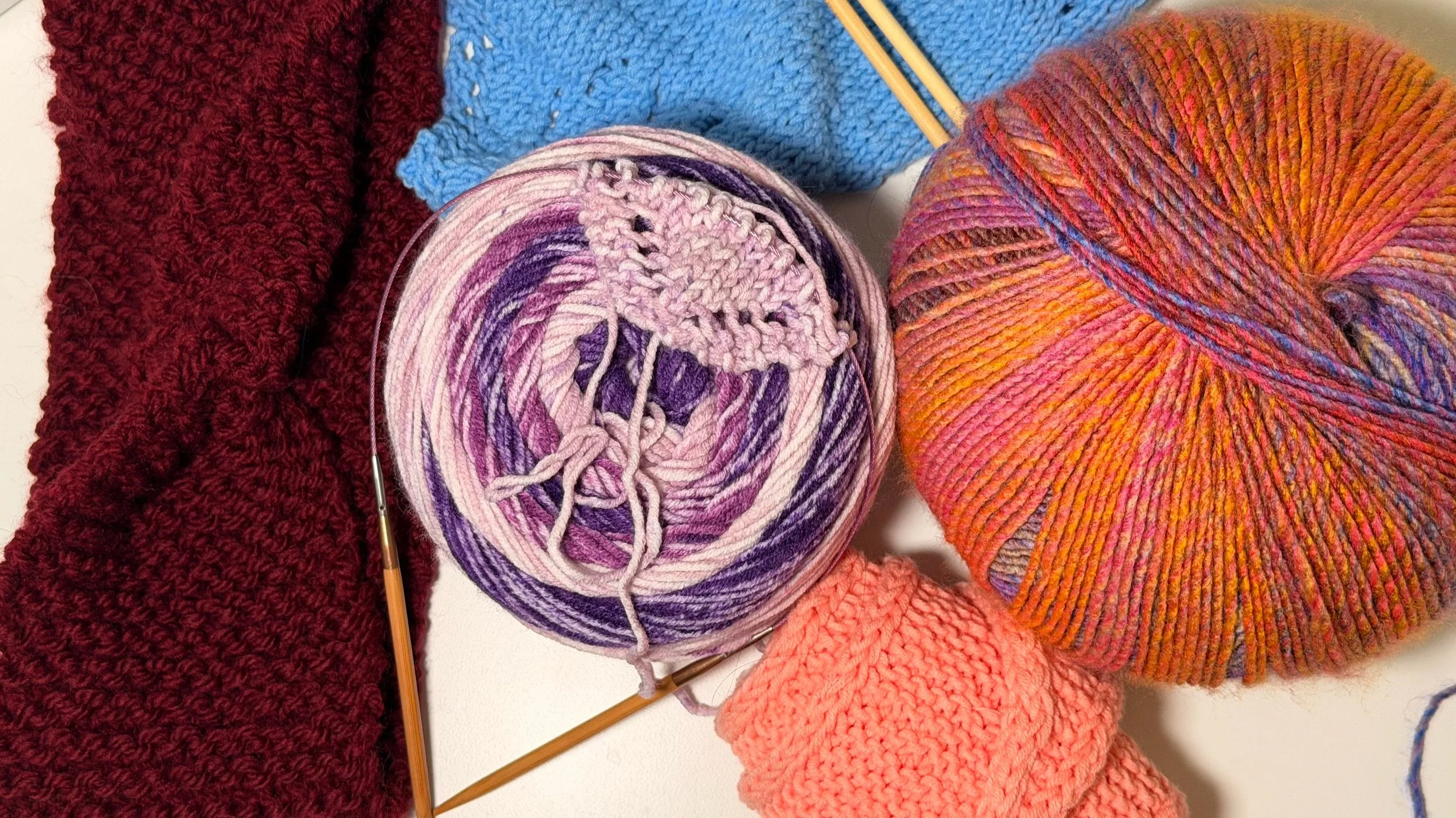 Various yarns and knitting projects including a red textured piece, a ball of multicolored yarn, a smaller knitted coral piece, and a ball of pink and purple yarn with knitting needles.