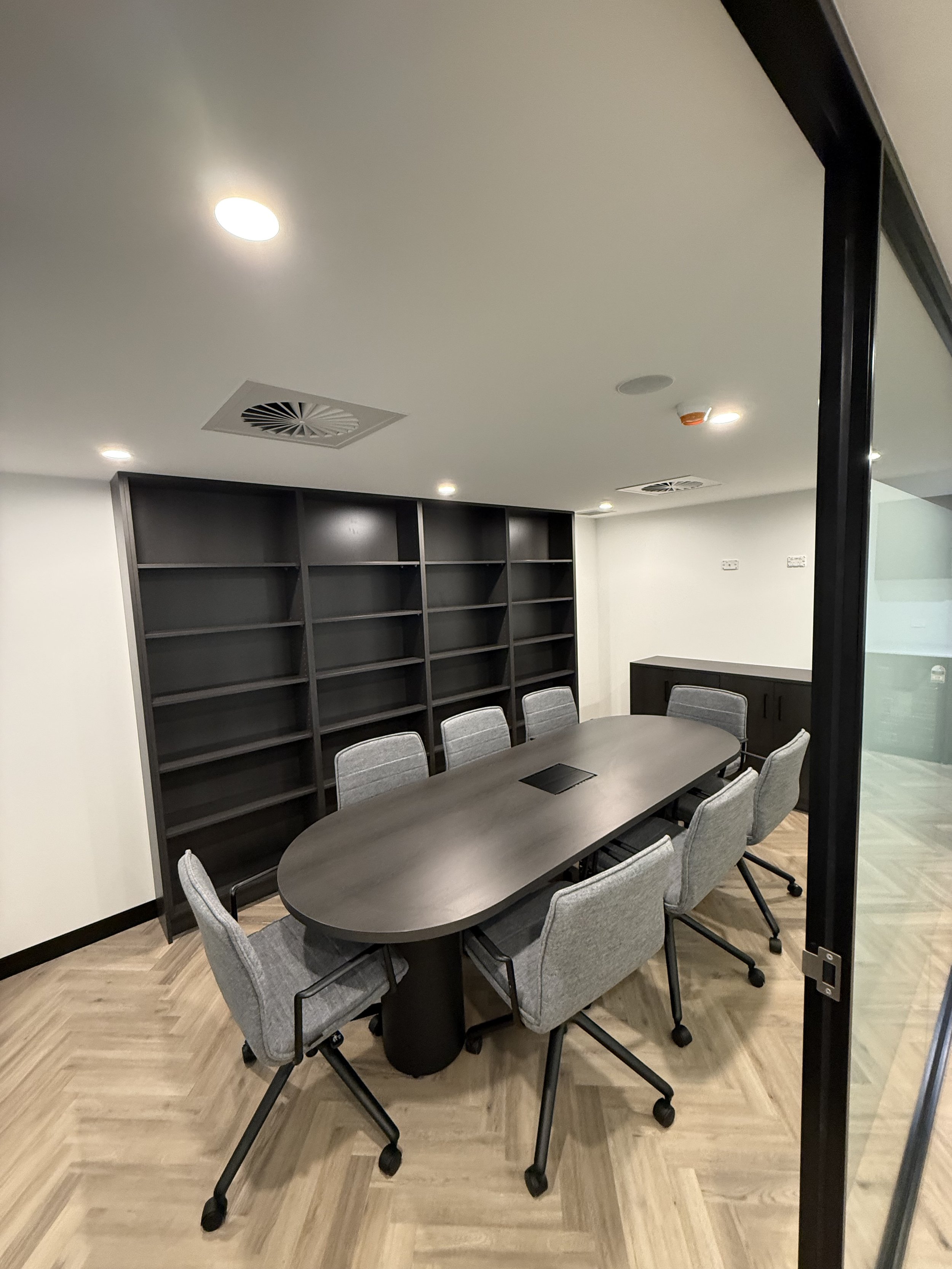 Empty modern conference room with a large oval table, eight gray chairs, a black bookshelf on the wall, and a small cabinet in the corner.