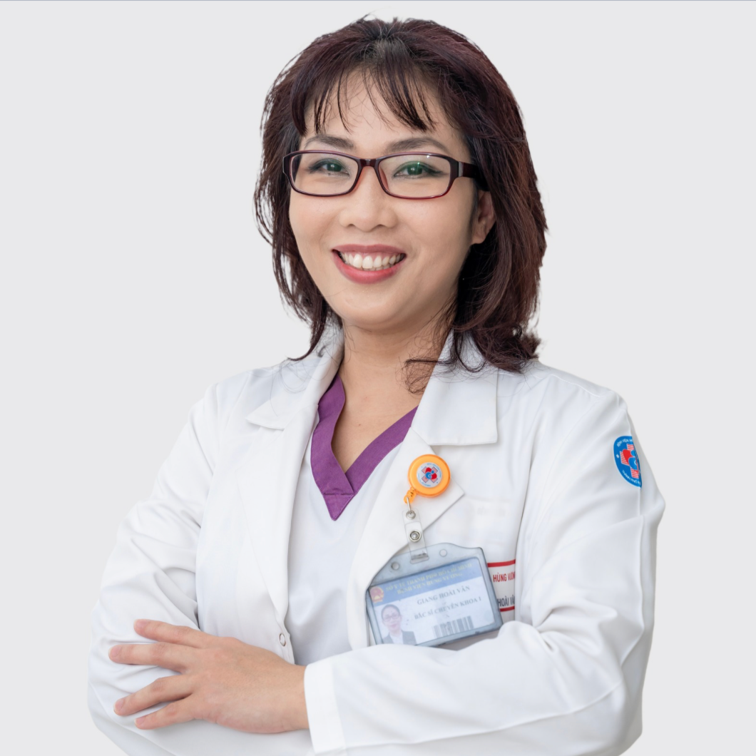 A female healthcare professional wearing glasses, a white lab coat with a badge, and a purple scrubs top underneath, smiling with arms crossed against a plain background.