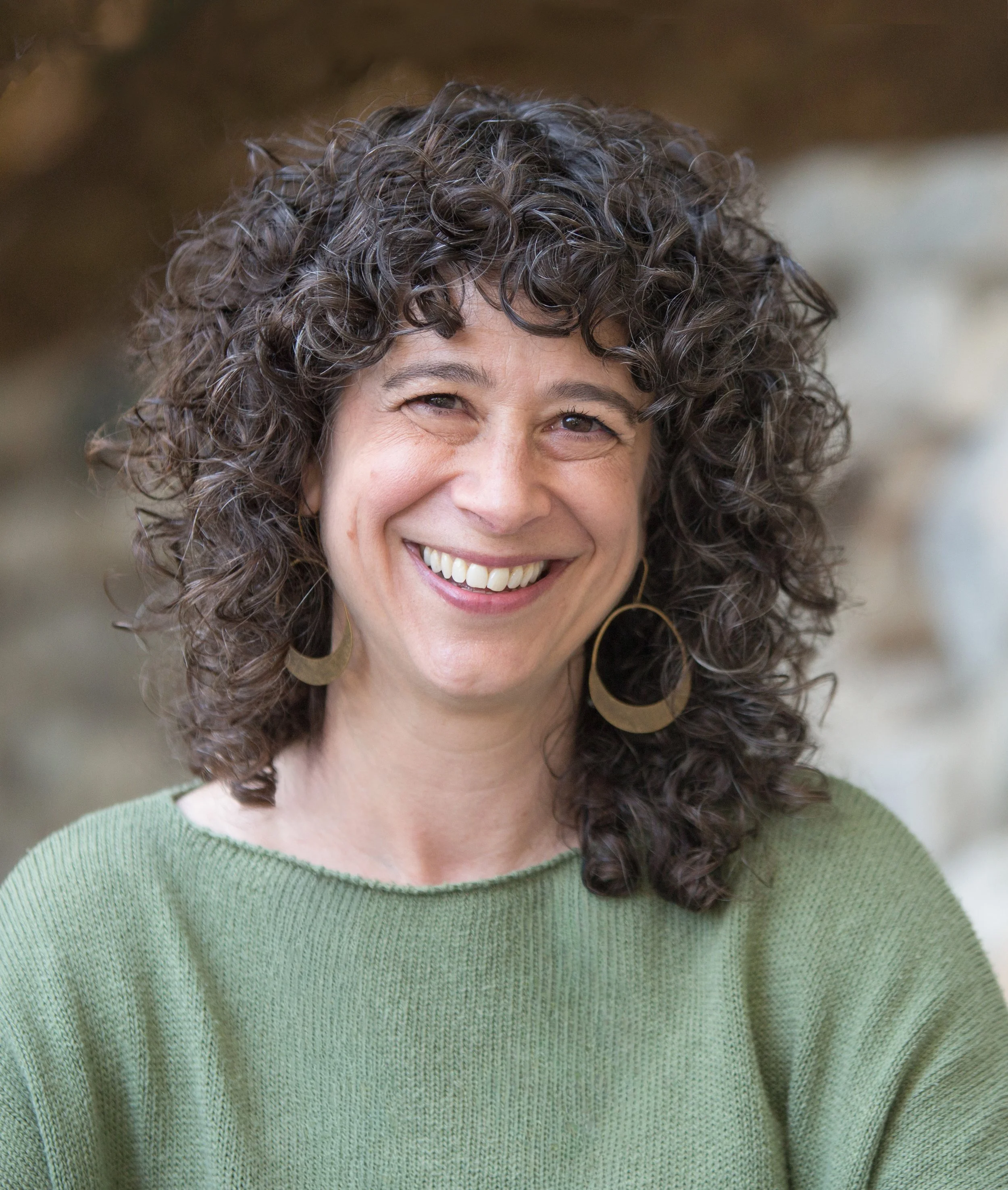 Head-and shoulders portrait of Shaleece, a white woman in her 50s with curly brown hair and wearing brass hoop earrings and a green knit top. She is smiling.