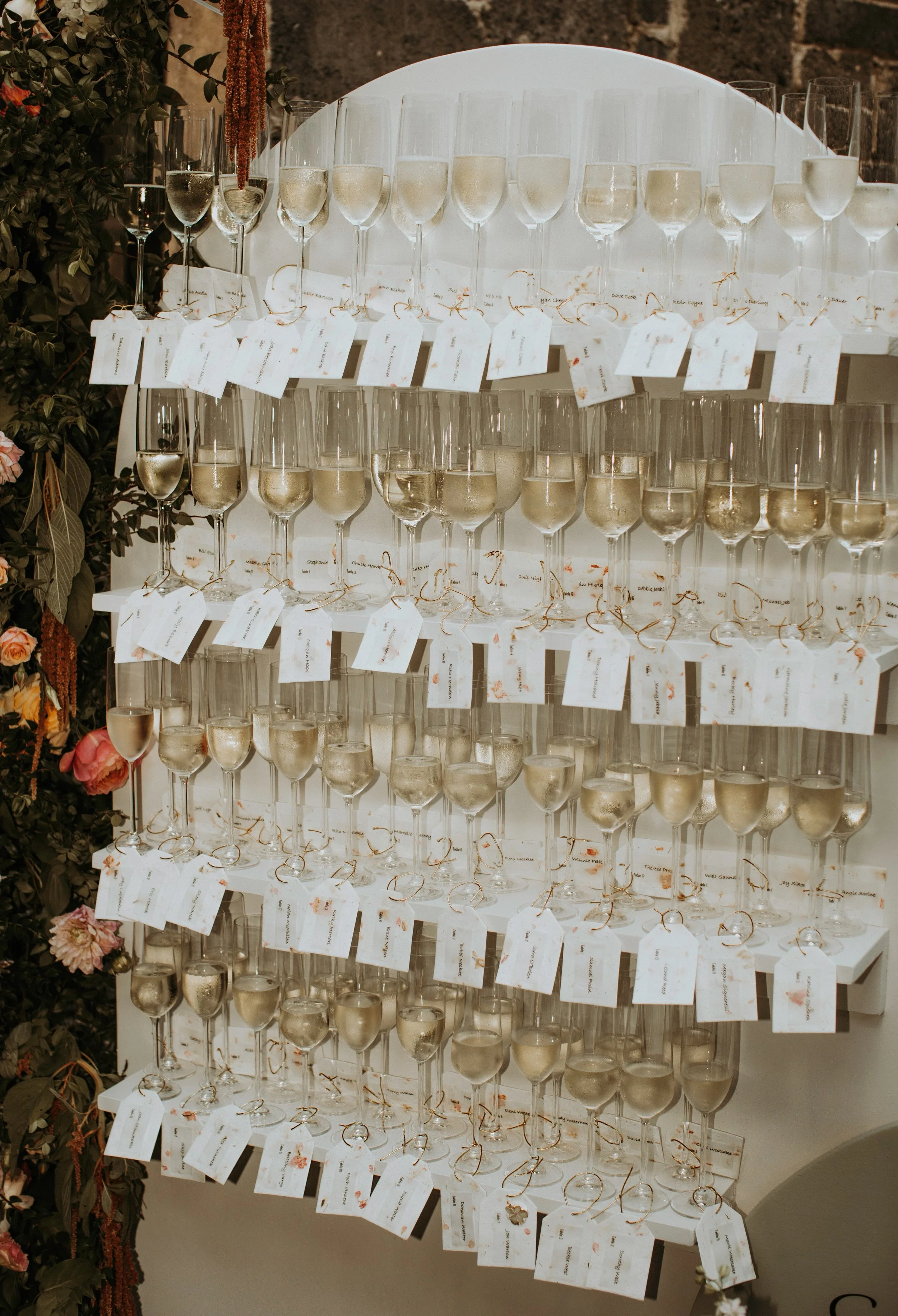 Display of multiple champagne glasses on a shelf, each with name tags attached, surrounded by floral decorations. Champagne wall seating chart or escort display for a wedding.