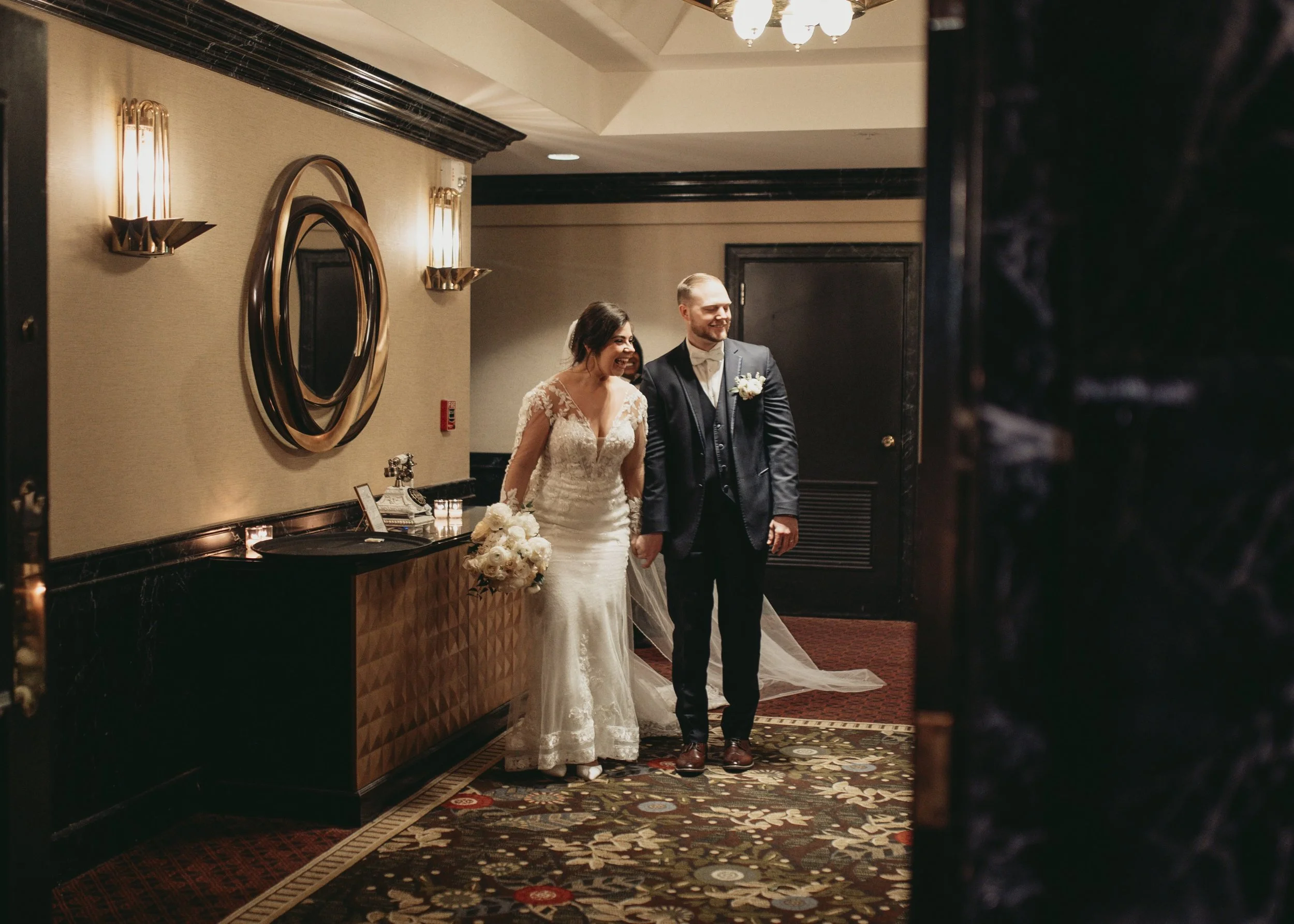 Bride and groom standing in a hallway with decorative mirrors and lights, holding hands, bride wearing a white gown holding a bouquet.