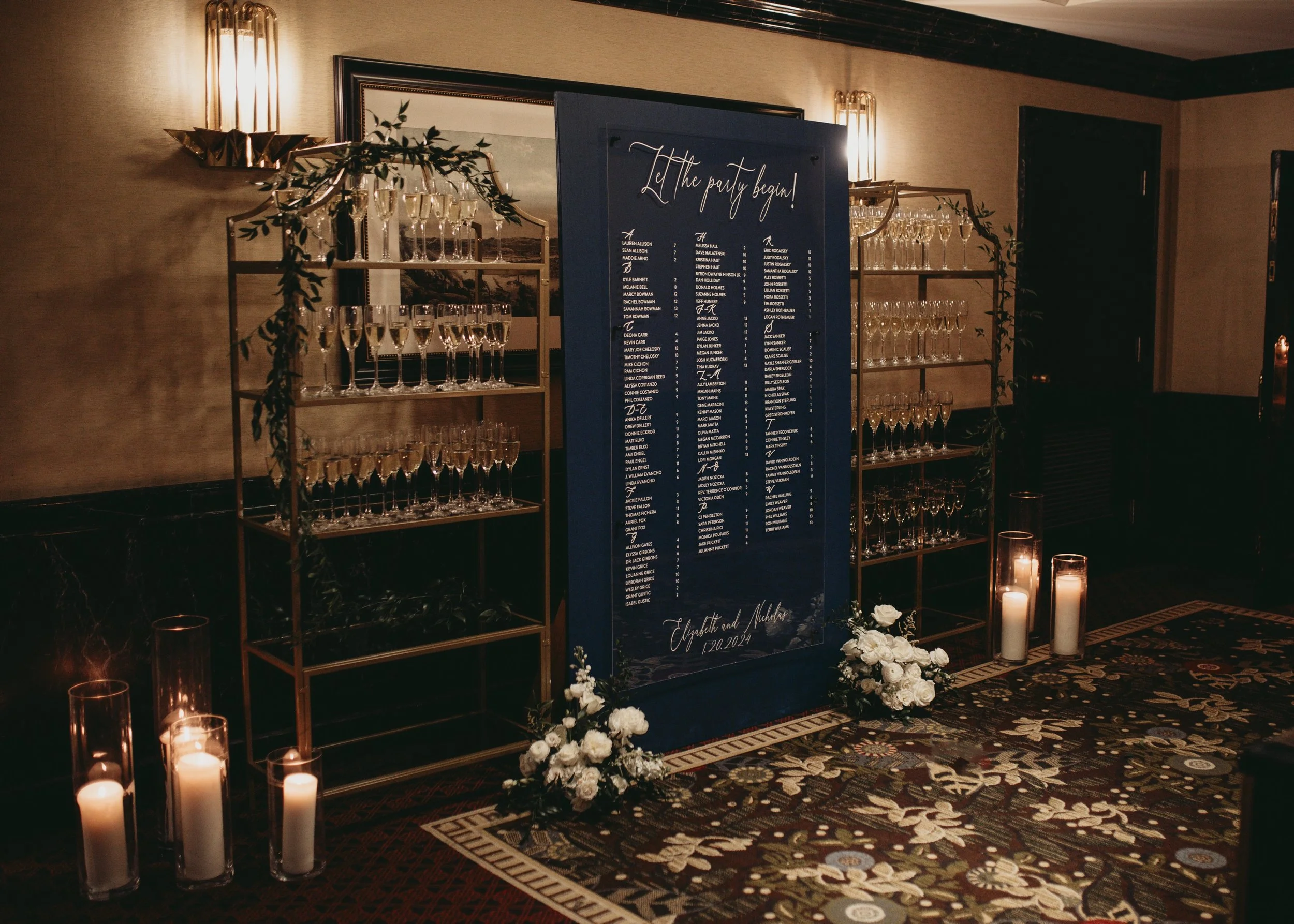 Wedding reception entrance with champagne glasses on shelves, a seating chart board, candles, and floral arrangements.
