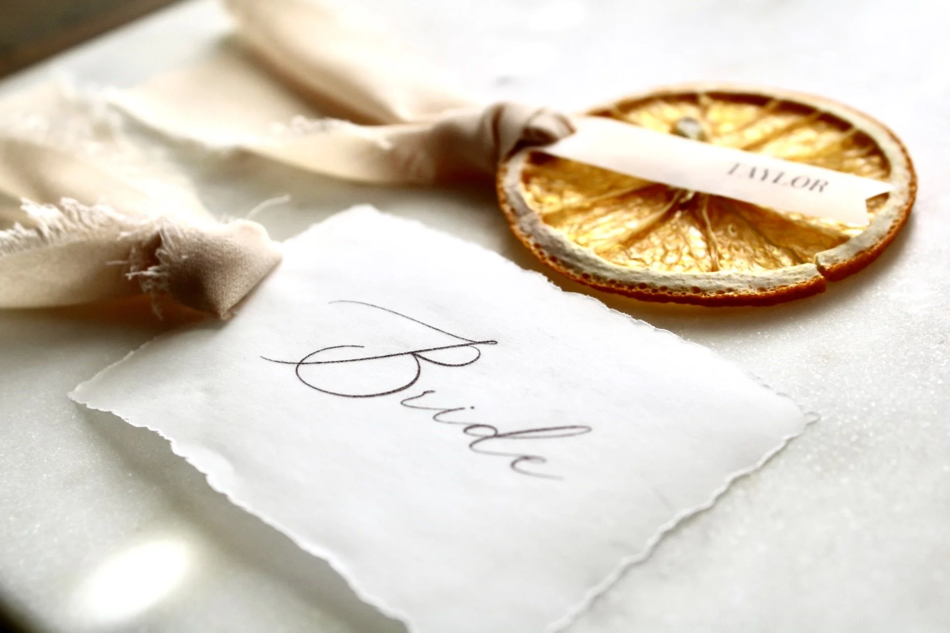 Close-up of a decorative wedding place card with the word 'Bride,' tied with beige ribbon and a dried orange slice.