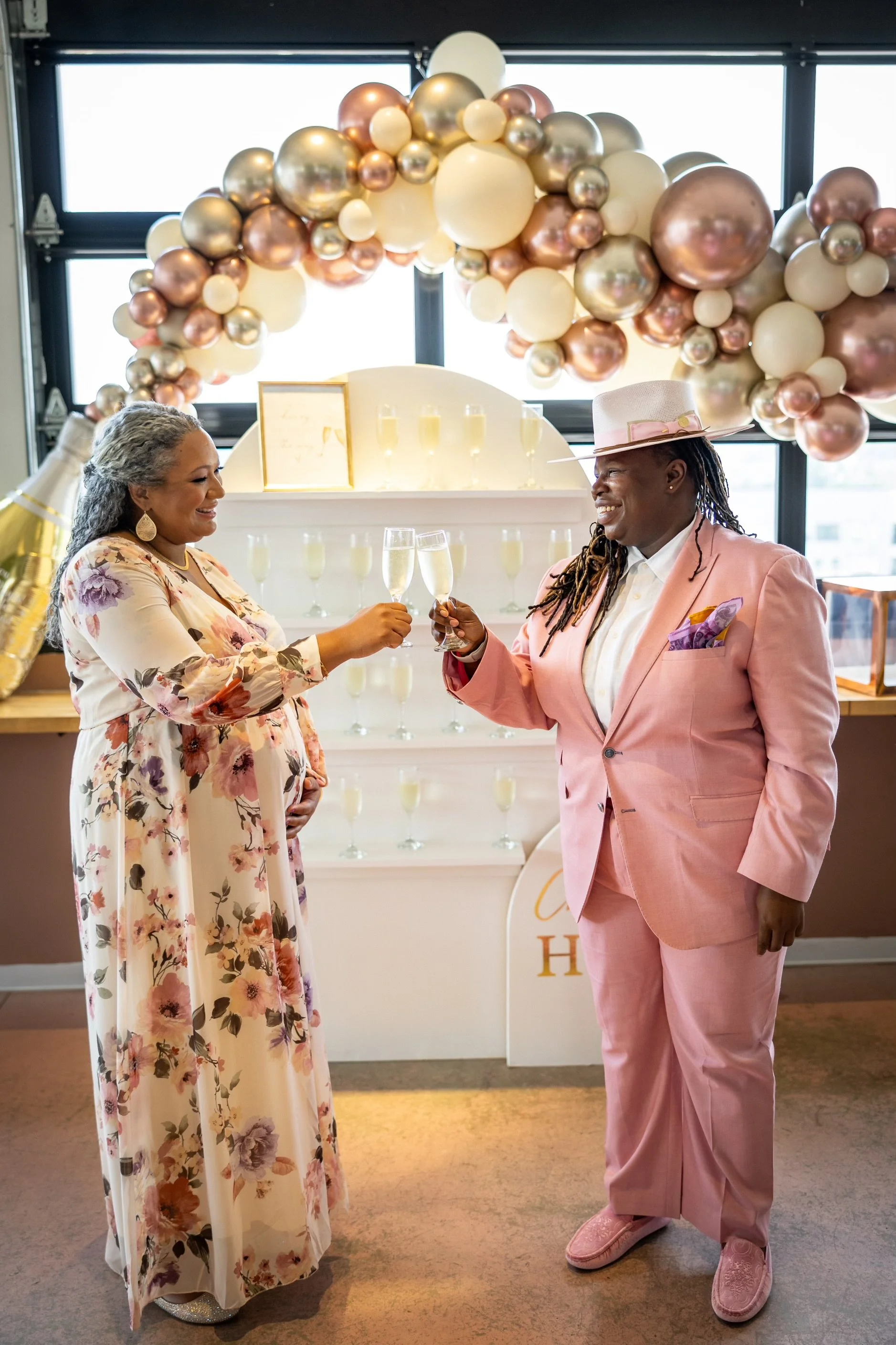 Two people clinking glasses in front of a decorative balloon arch.