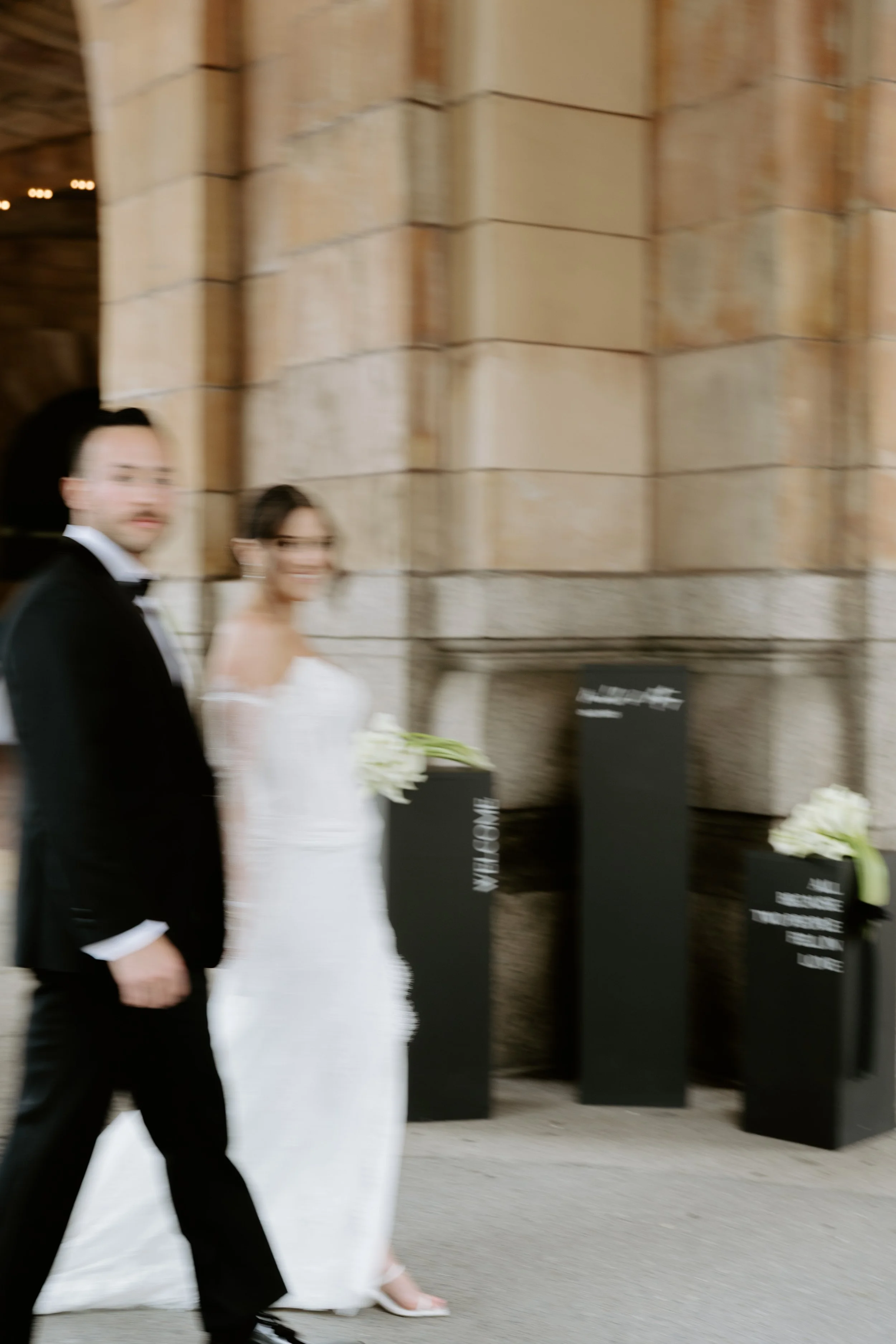 Bride and groom standing in a decorated hallway, the bride wearing a white gown holding a bouquet, and the groom in a dark suit with a boutonniere.