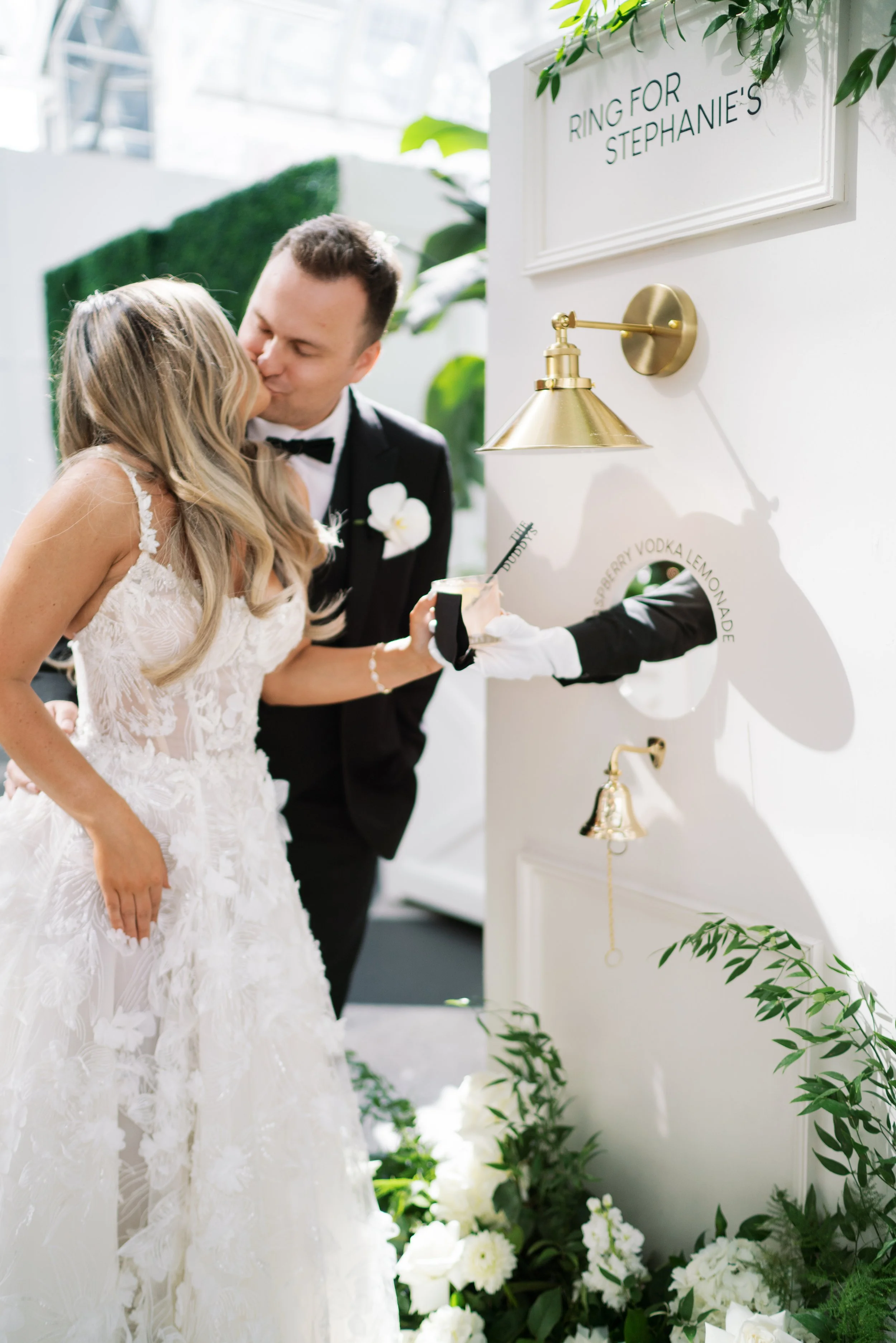Bride and groom at wedding reception, with a drink service on the wall labeled 'Ring for Stephanie's' surrounded by white flowers and greenery. Living cocktail or champagne wall and seating chart.
