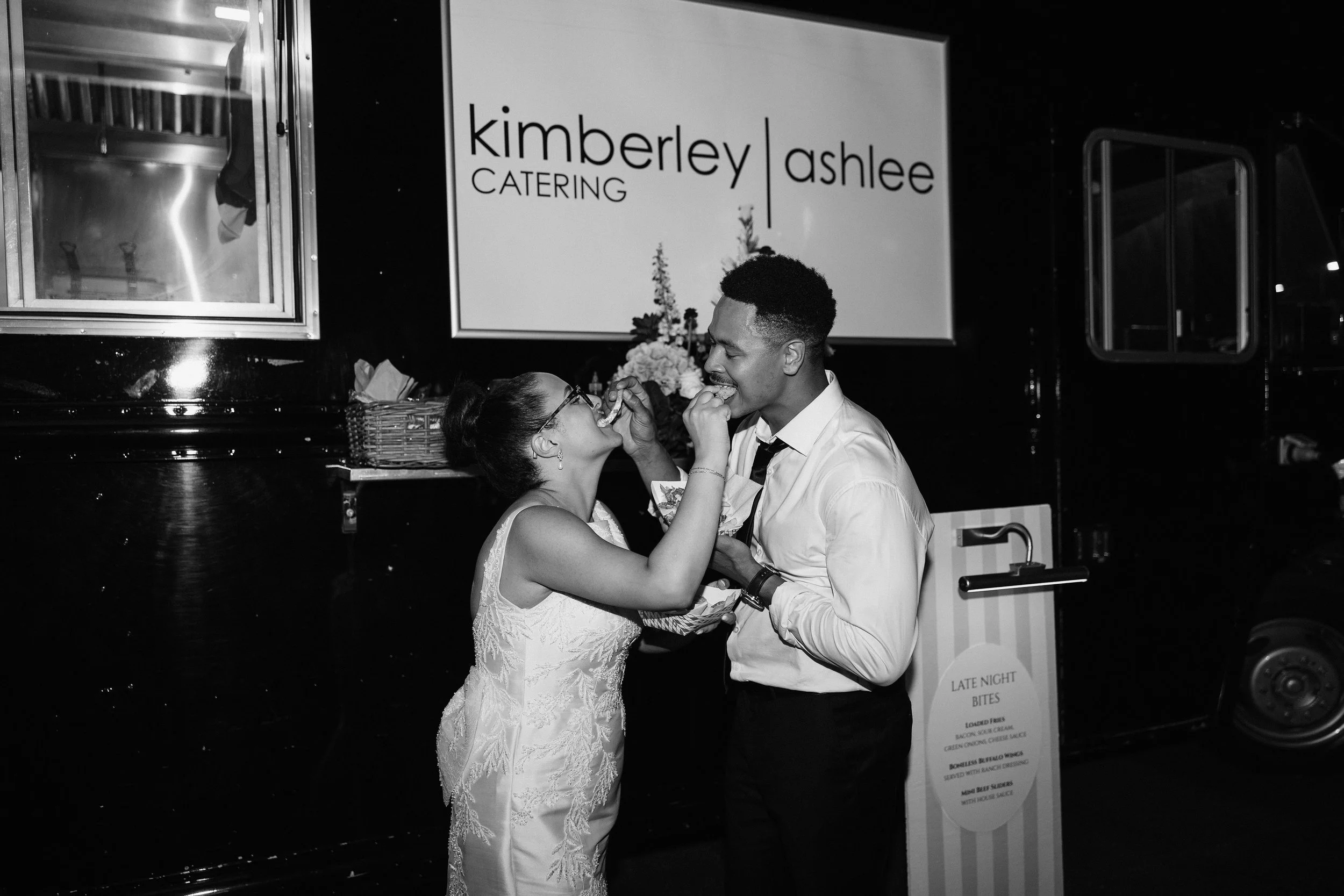 Bride and groom holding hands in a hotel hallway, decorated with elegant mirrors, wall sconces, dark wood moldings, and floral carpet; bride holding a bouquet of white flowers.