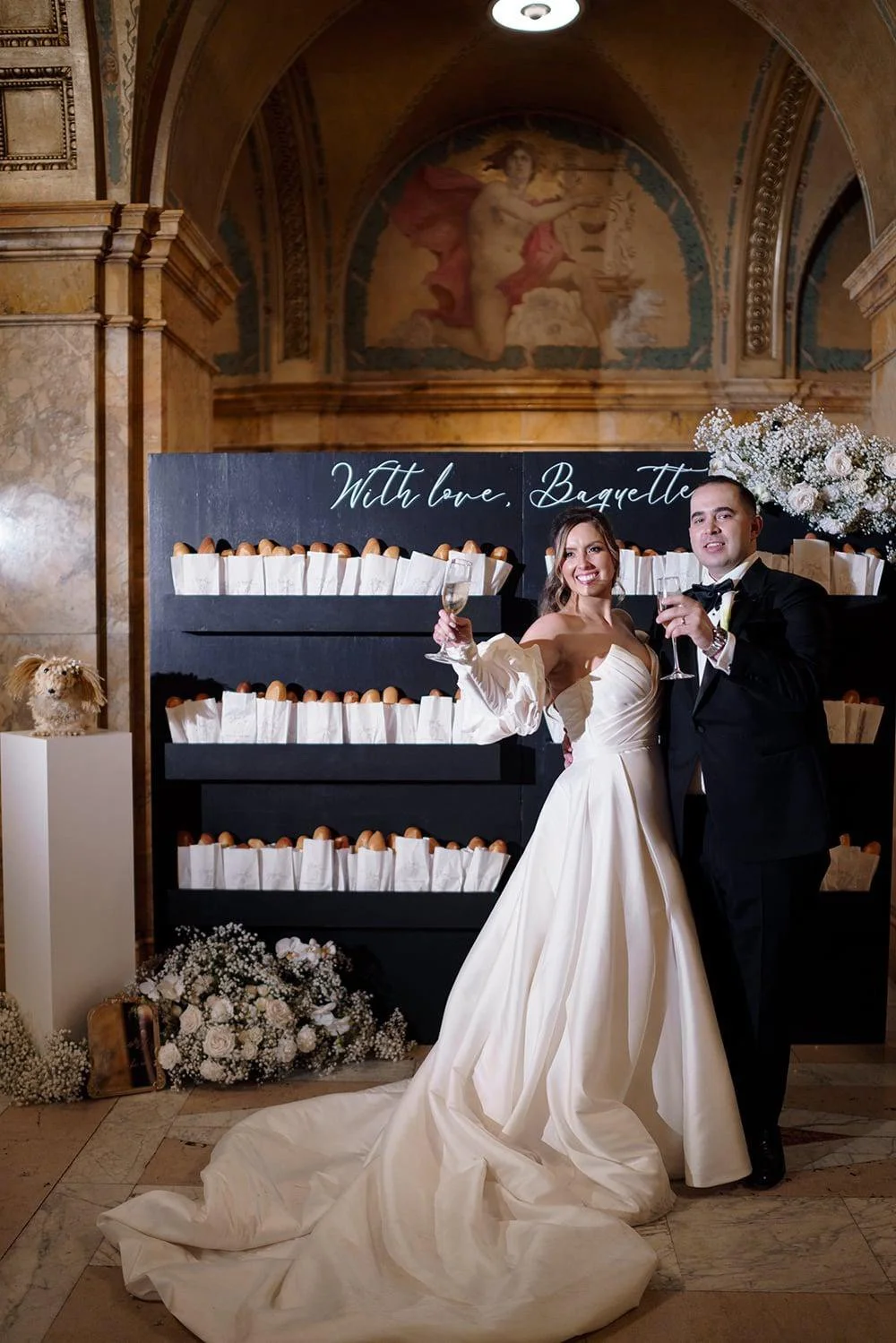 Display of multiple champagne glasses on a shelf, each with name tags attached, surrounded by floral decorations. Champagne wall seating chart or escort display for a wedding. 