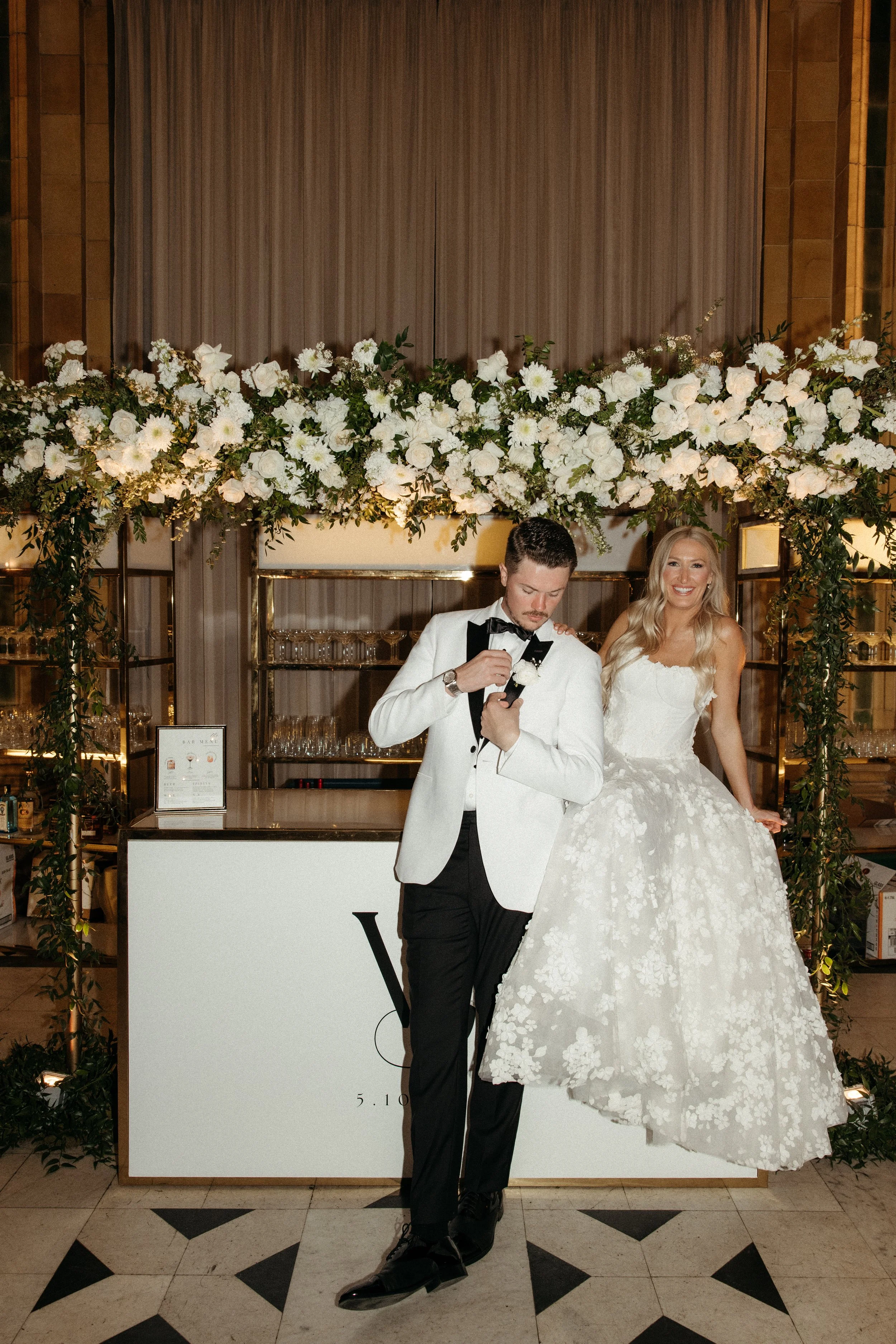 Two people toast with champagne glasses at a celebration, standing in front of a champagne wall with glasses. One wears a floral dress, the other a pink suit and hat. A balloon arch decorates the background.