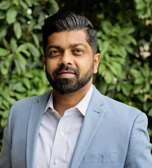 A man with dark hair and a beard wearing a light blue suit jacket and a white checked shirt, standing outdoors with green foliage in the background.