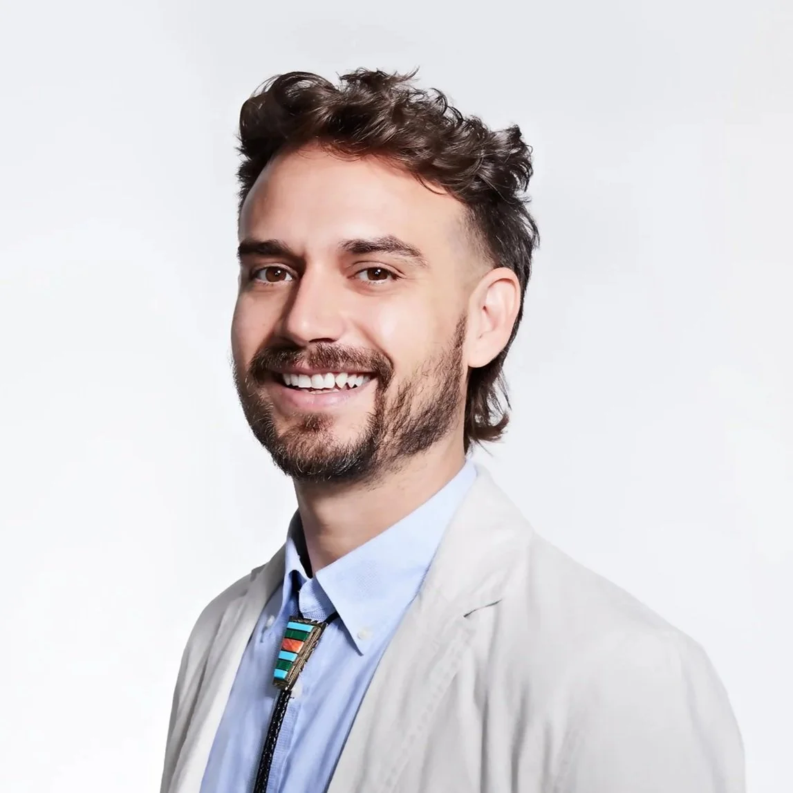 Portrait of a man with dark, wavy hair and a beard, smiling, wearing a light-colored blazer over a blue shirt with a decorative zipper, against a plain white background.