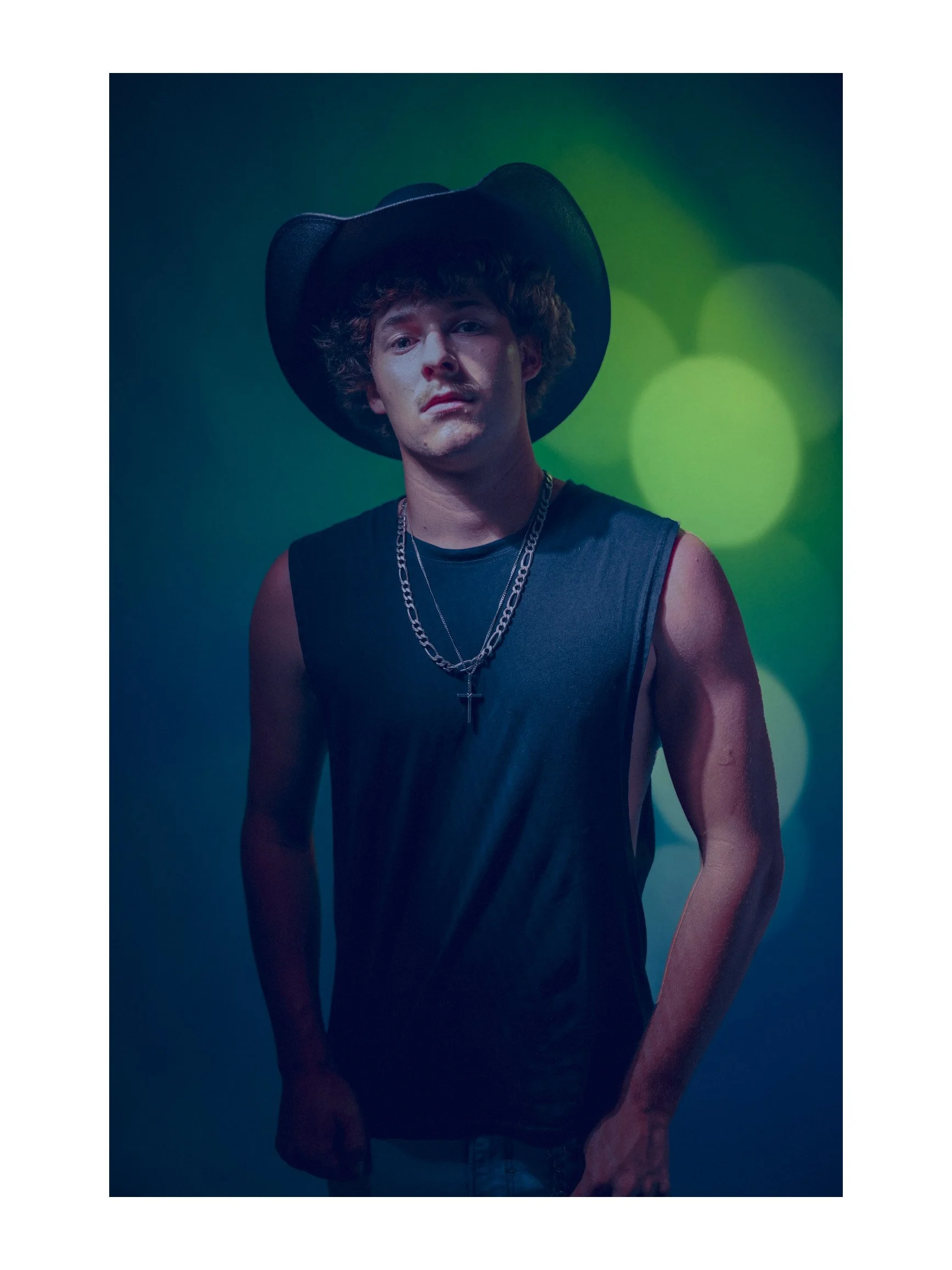 Young man with curly hair wearing a black sleeveless shirt, layered chains with a cross pendant, and a wide-brimmed black hat, standing against a colorful, blurred background with green and blue tones.
