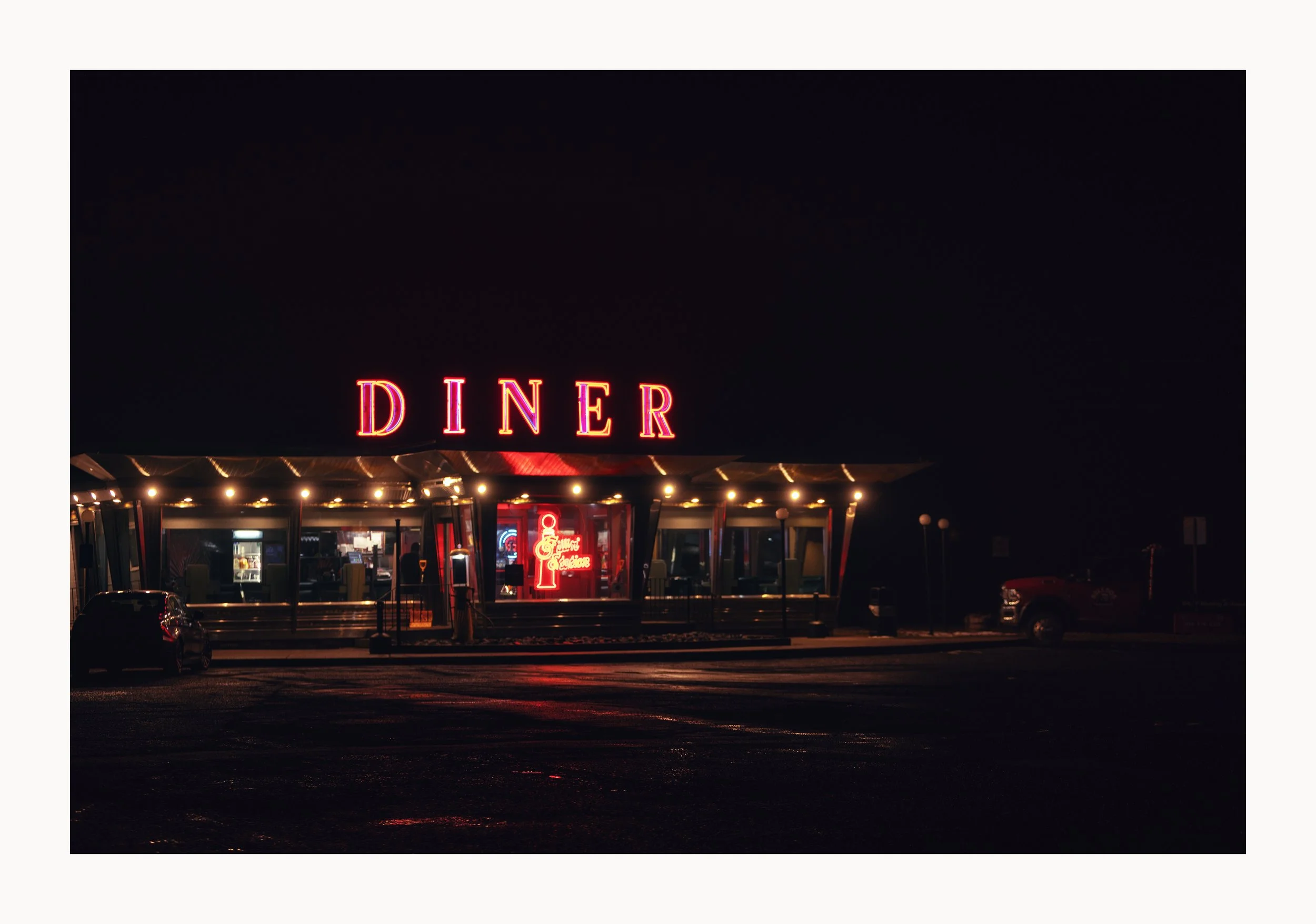 Nighttime view of a vintage-style diner with neon lights, including a large illuminated 'DINER' sign, and a neon ice cream cone sign in the window.