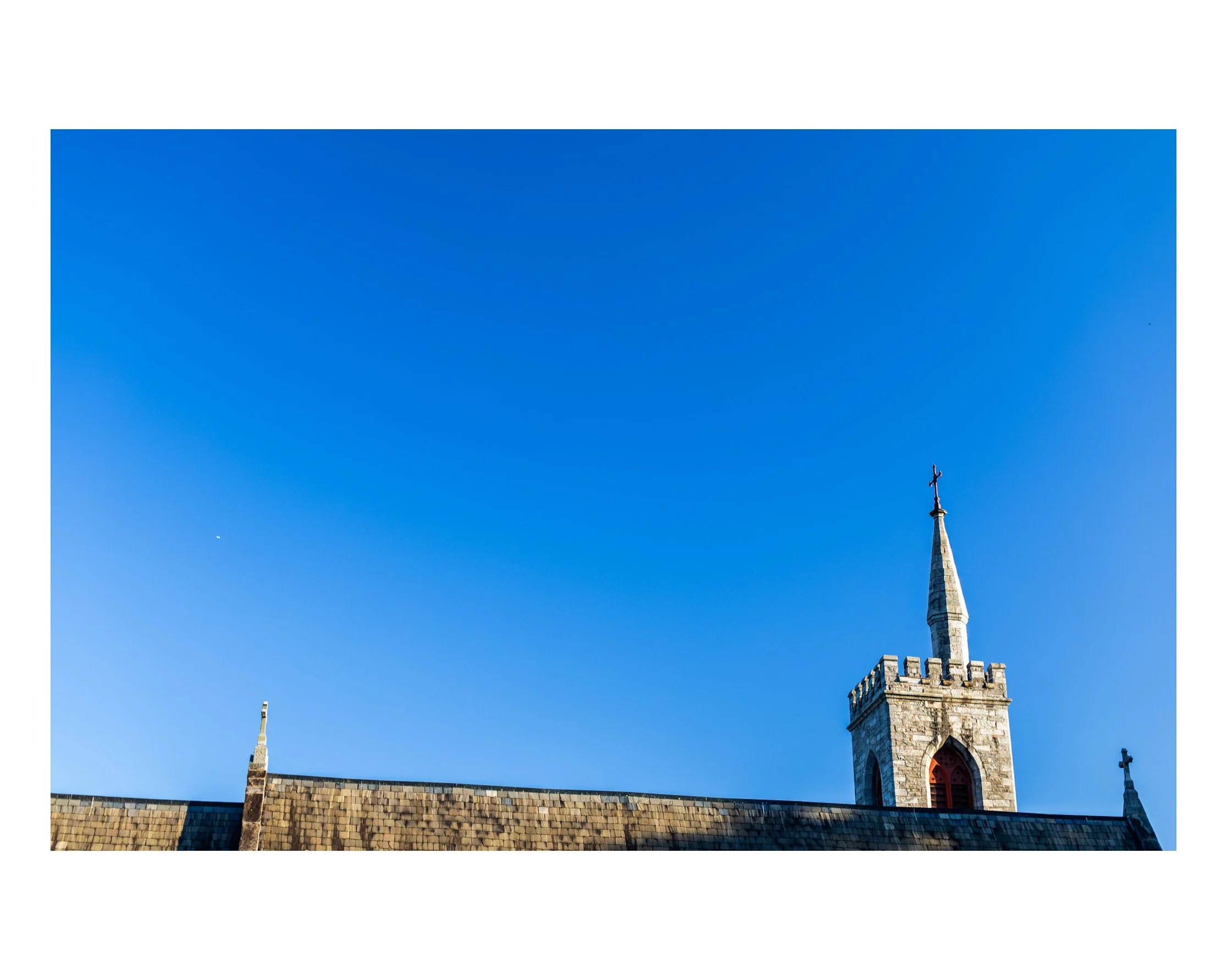 A church steeple with a cross on top against a clear blue sky, with part of the roof visible in the foreground.