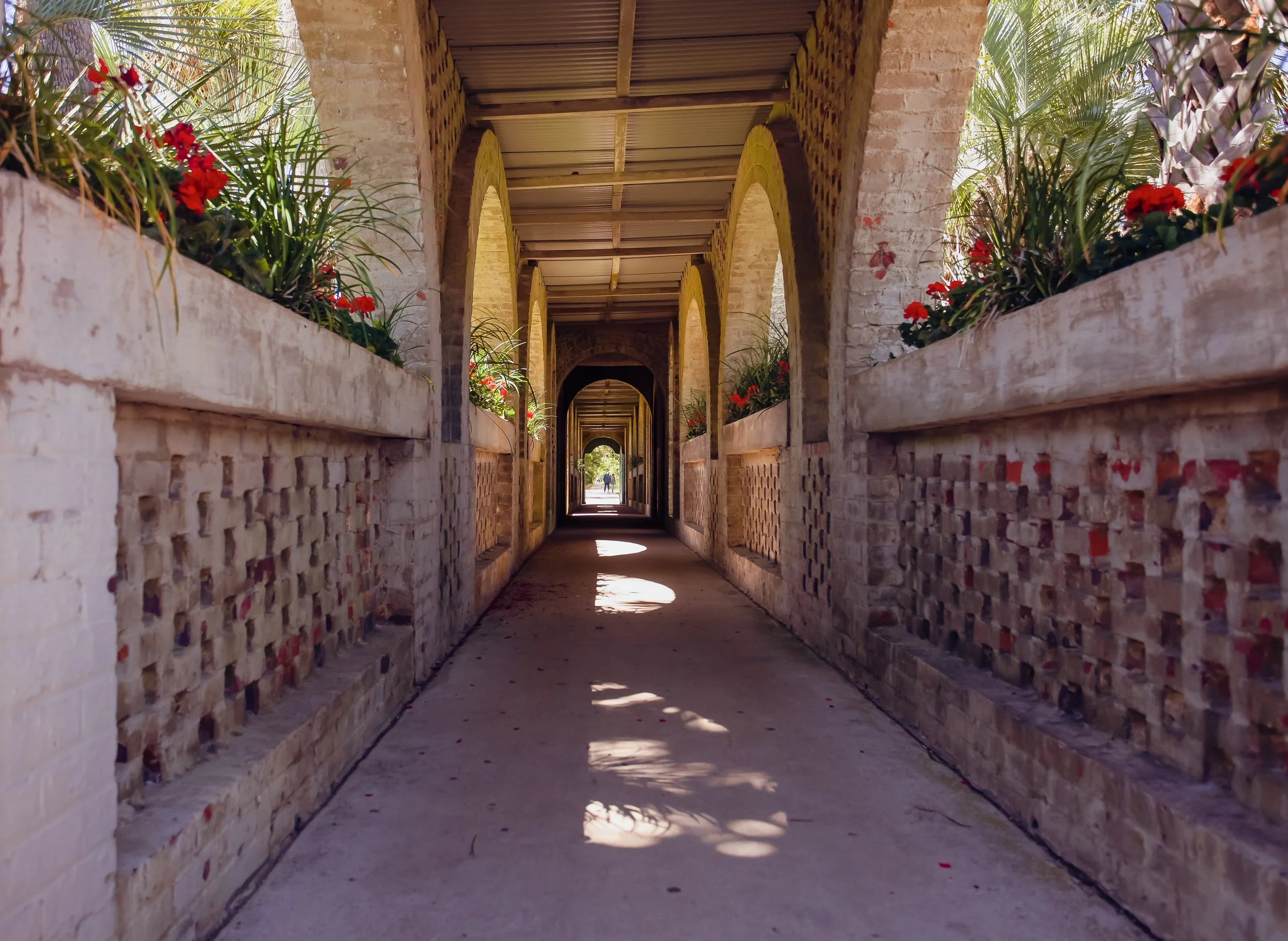 A covered stone walkway with arches, brick walls, and flower boxes with red flowers along the sides. Sunlight creates shadows on the ground, and there are people visible in the distance.