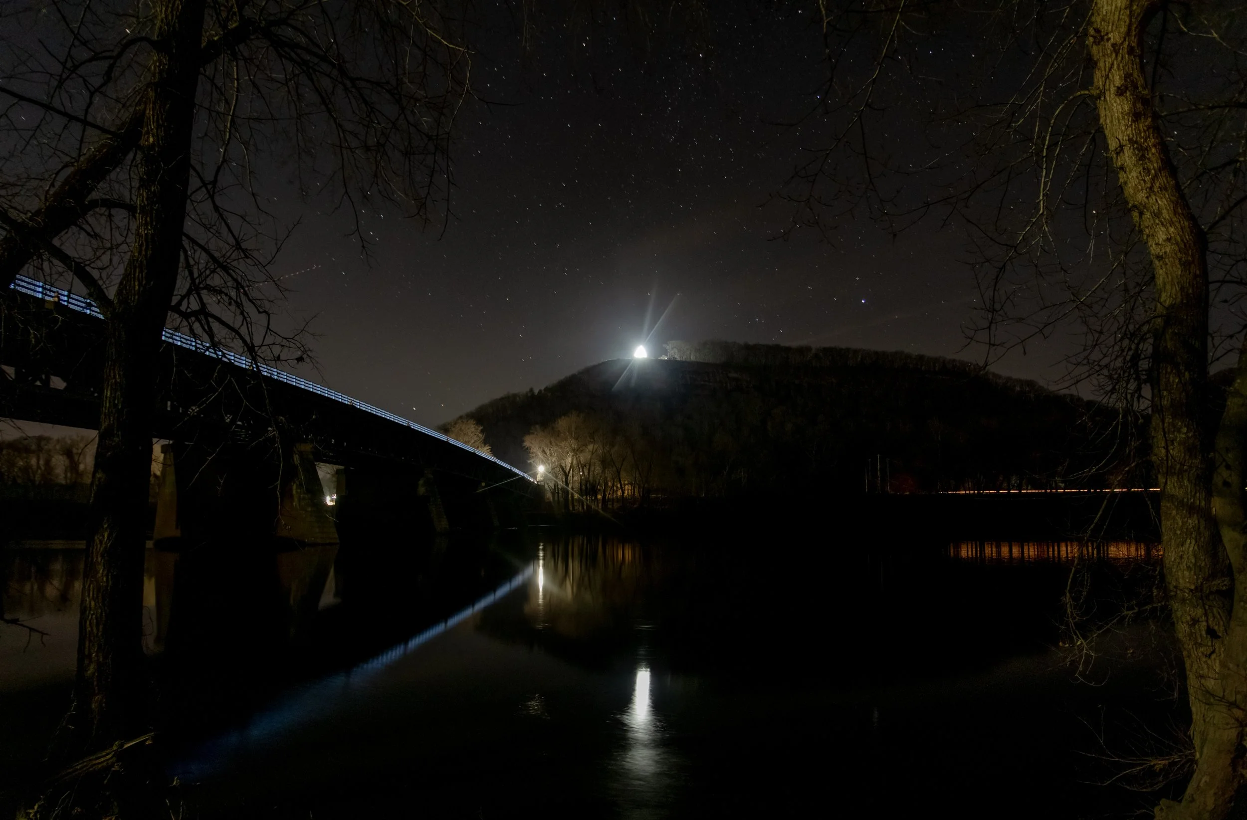 Nighttime scene with a bridge over a river, illuminated by artificial lights, with trees on both sides. A hill with a bright lighthouse or building with a light beam is visible in the background, under a starry sky.