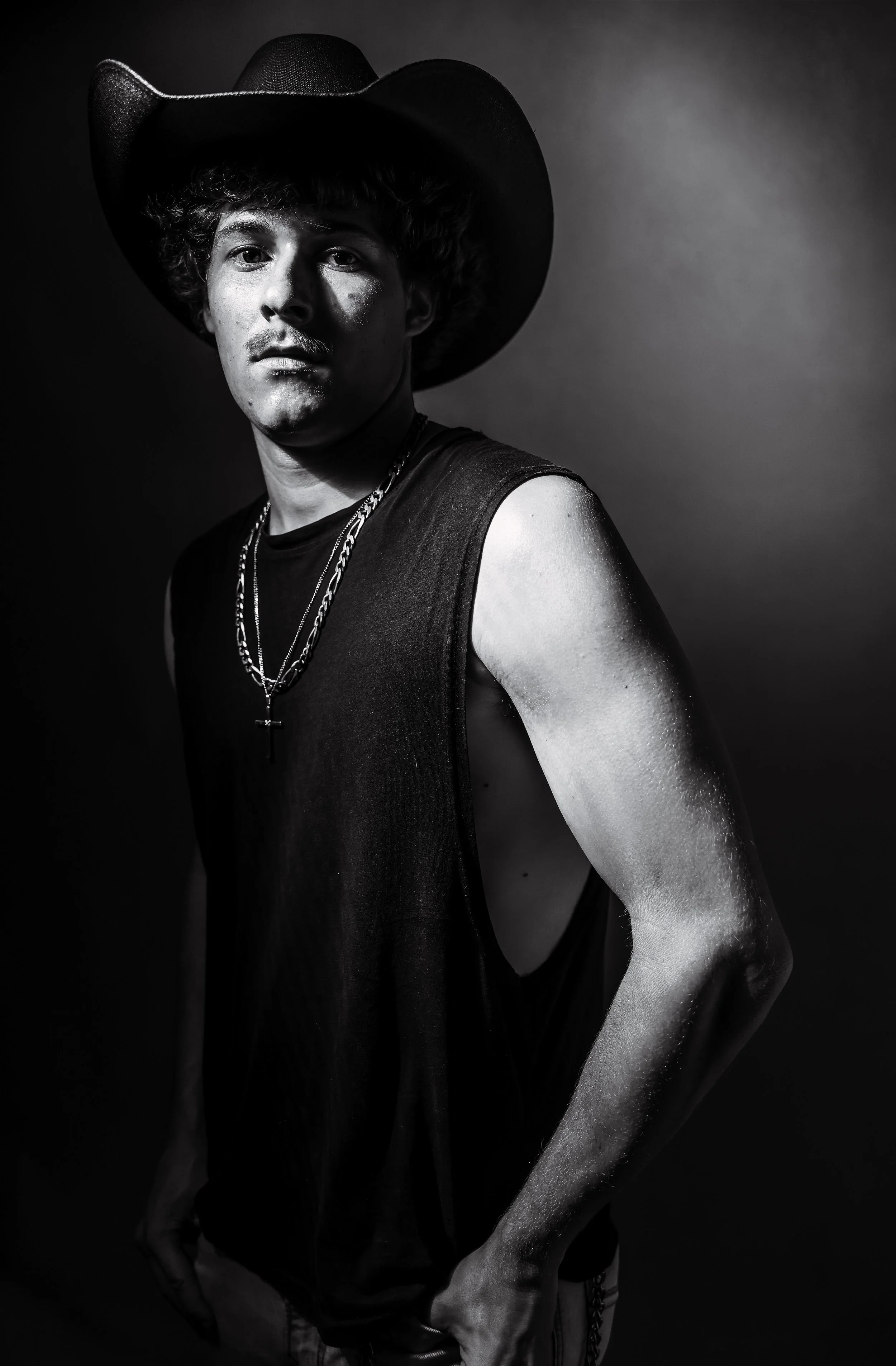 Black and white portrait of a young man wearing a cowboy hat, sleeveless shirt, and layered necklaces, posing with one hand on his hip against a plain background.