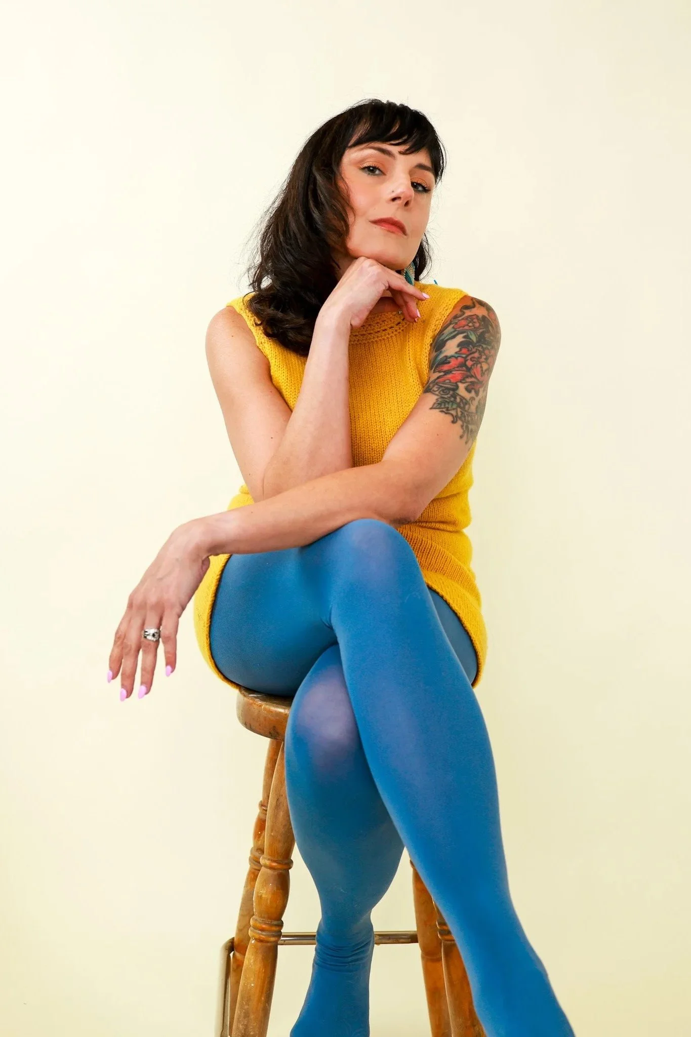 Portrait of a woman with dark wavy hair and a tattooed arm, sitting on a wooden chair, wearing a yellow sleeveless top and blue tights, against a plain yellow wall.