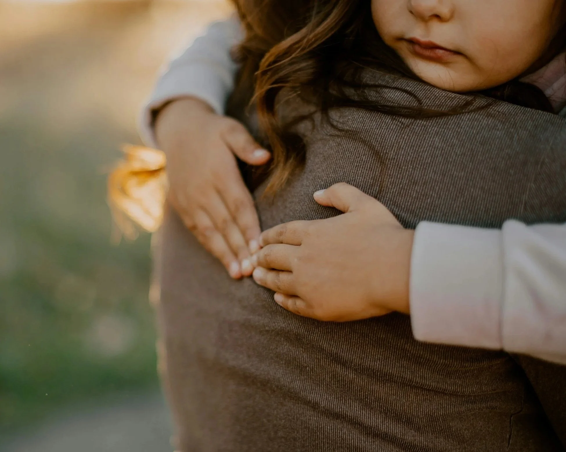 Close-up of a child's face hugging an adult, with their arms wrapped around each other, outdoors in warm lighting.