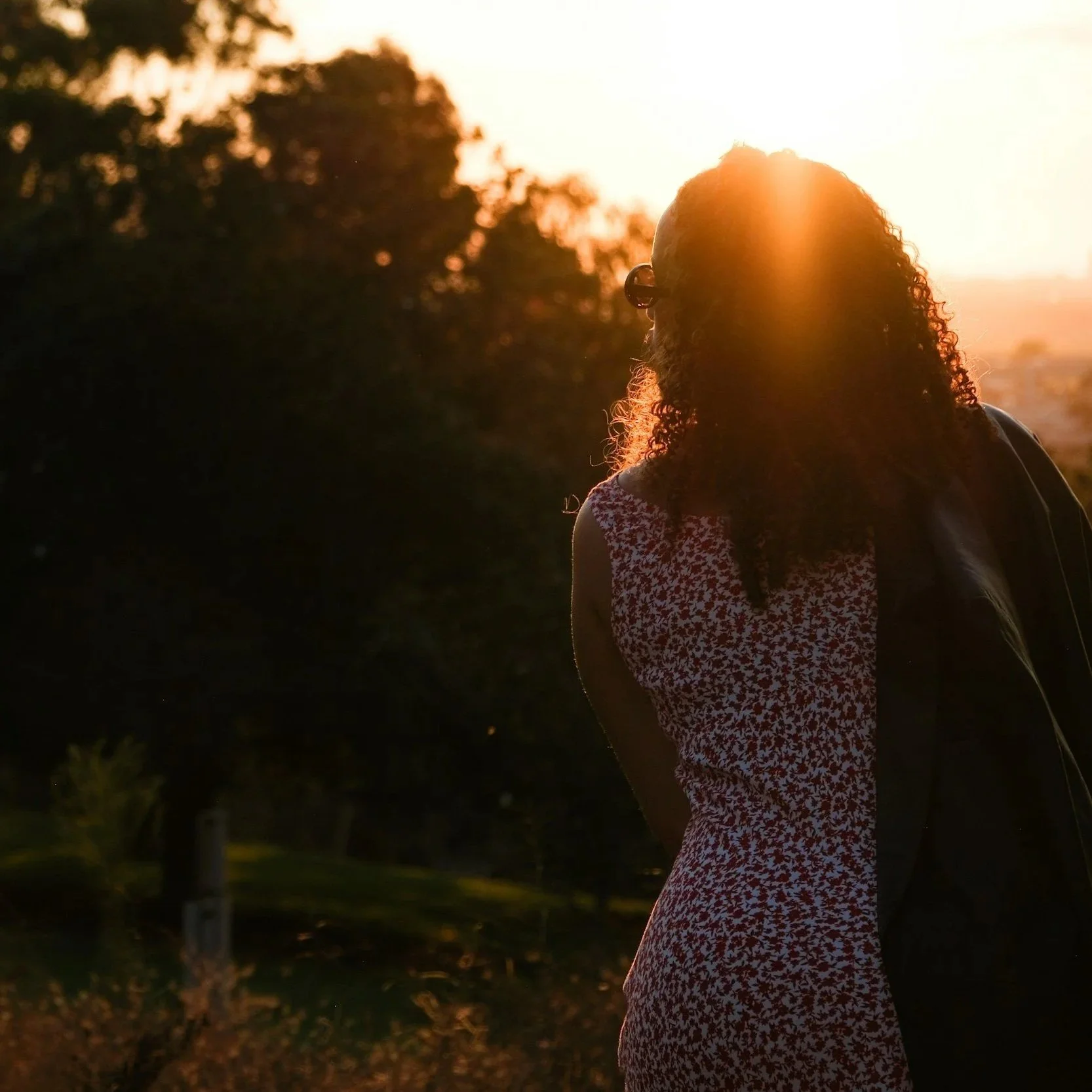 A woman with curly hair and glasses standing outdoors at sunset, wearing a red and white patterned dress.