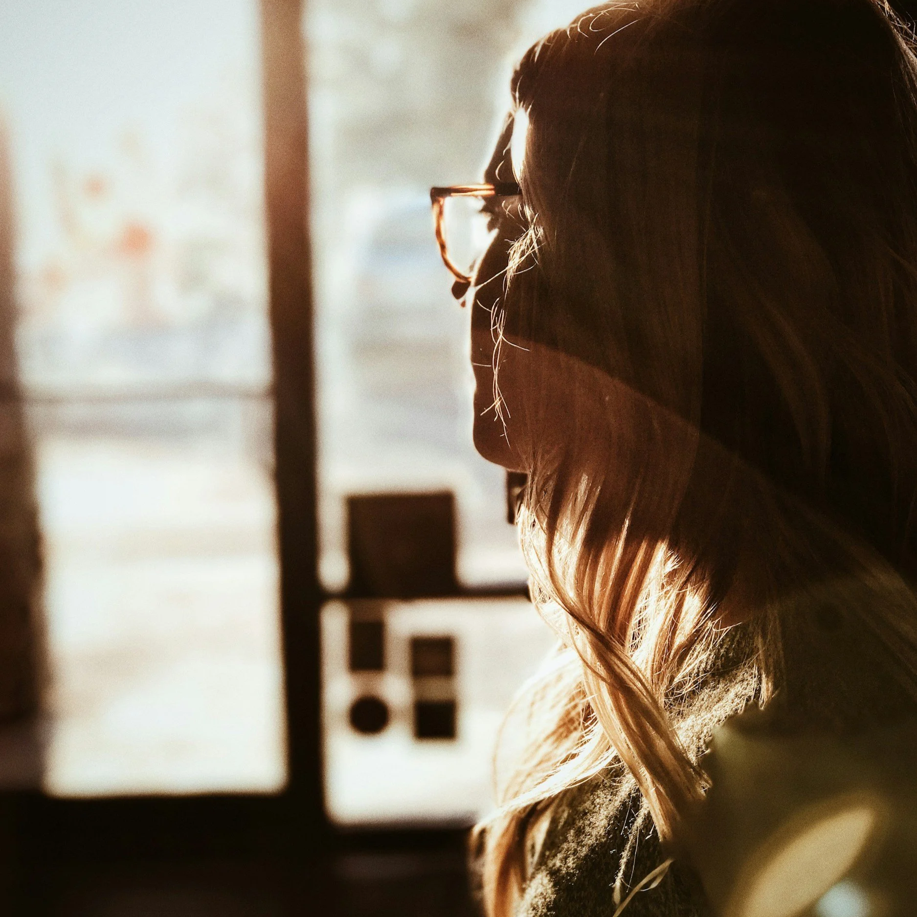 Close-up of a person's face with glasses and long hair illuminated by warm sunlight, sitting by a window with blurred background.