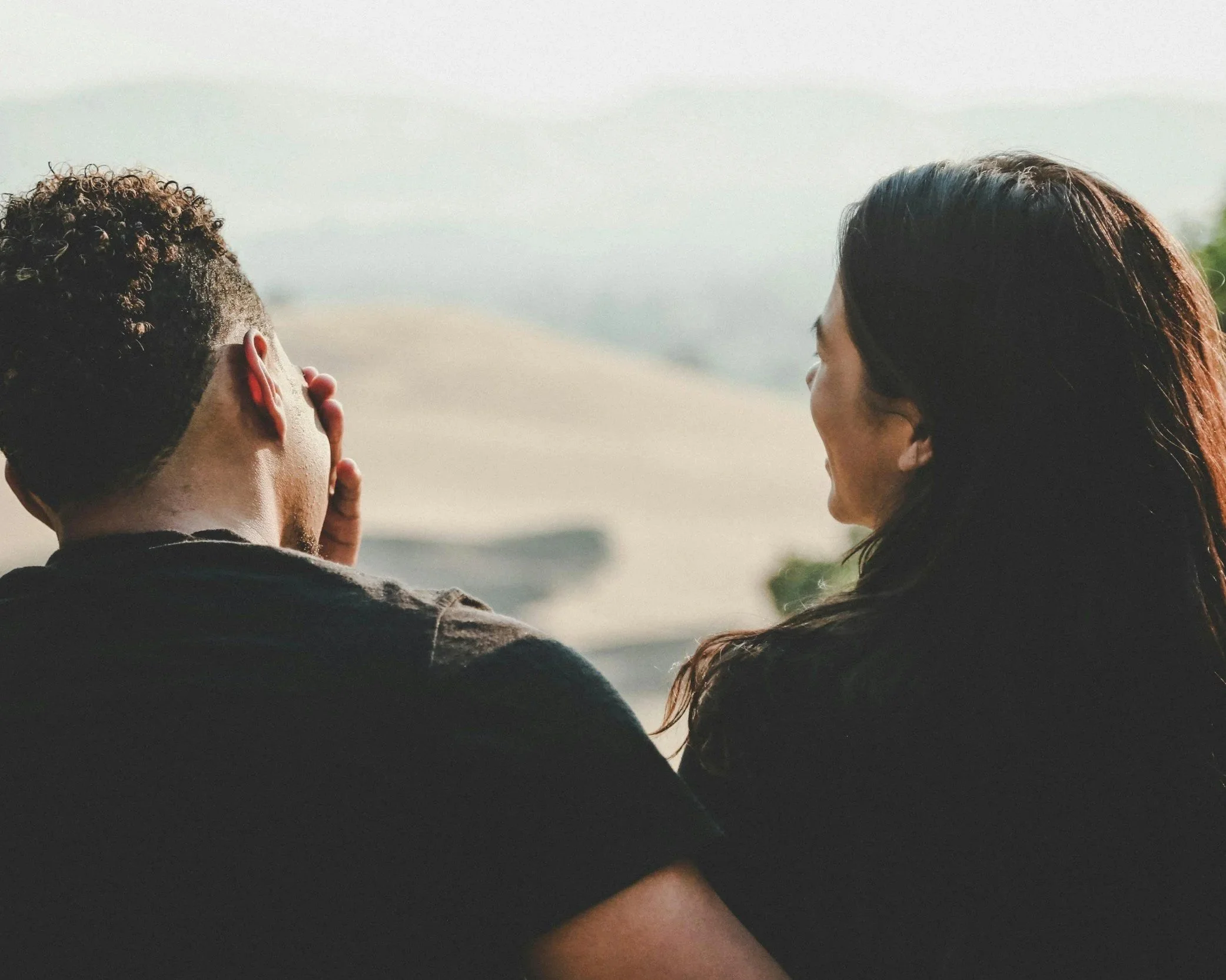 The image shows the back view of a young man with curly hair and a woman with long straight hair, both looking out at a scenic landscape with water and distant hills during daytime.