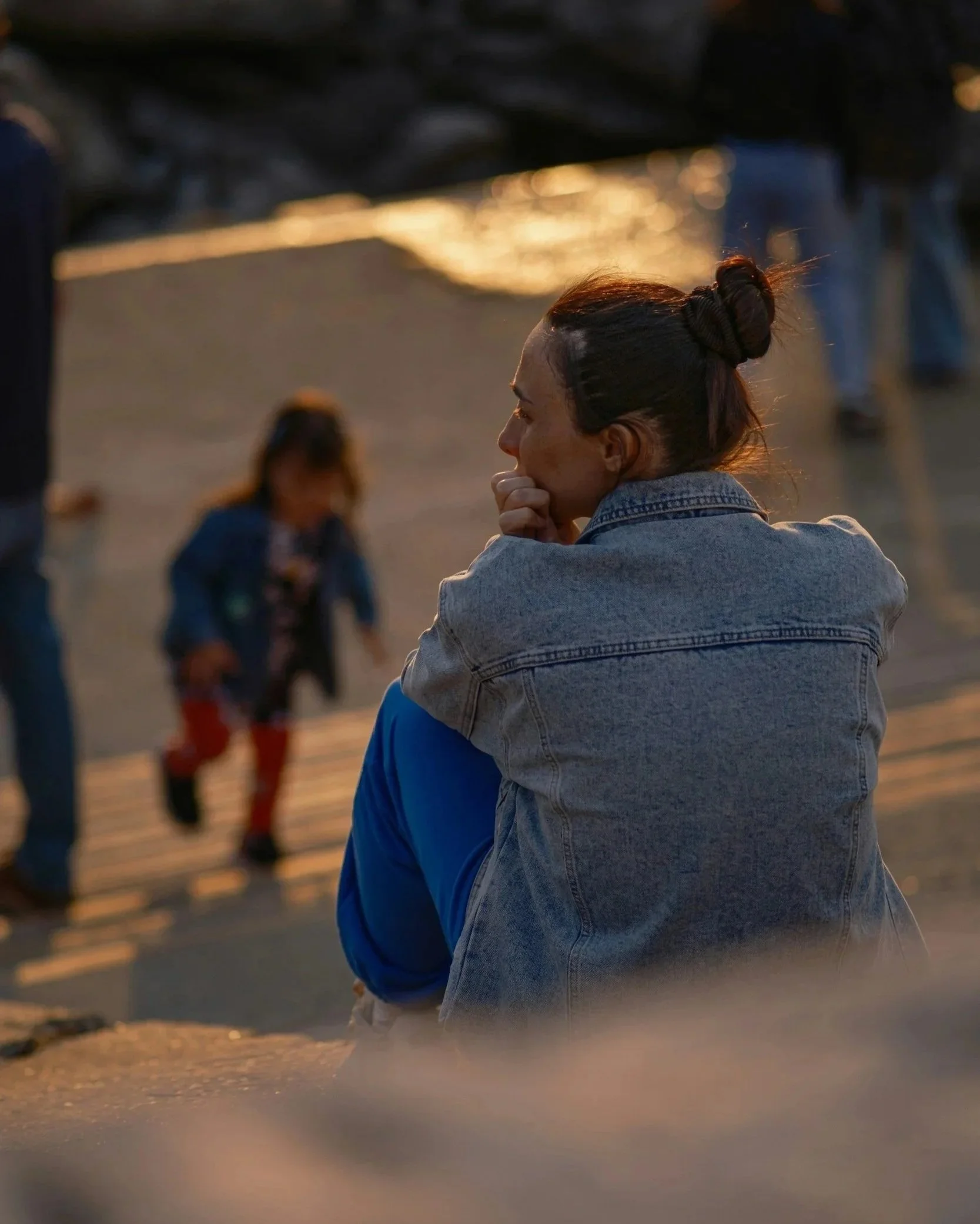 A woman with dark hair in a bun and wearing a denim jacket is sitting by the street at sunset, with blurred pedestrians in the background.